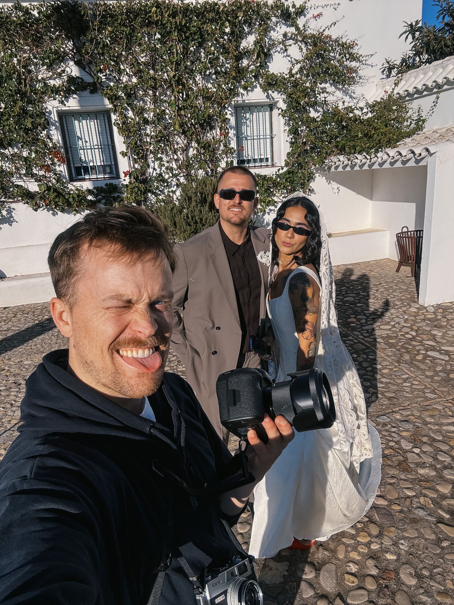 Dylan of Miles north Creative . Co takes selfie with camera while couple in wedding attire poses behind in sunny courtyard.