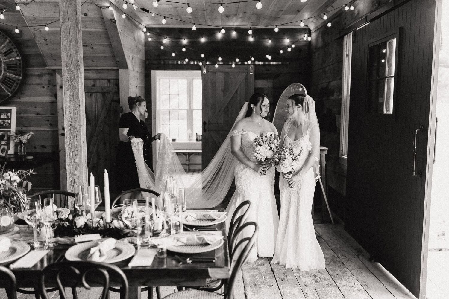 Two brides in wedding gowns share a moment inside a rustic barn venue with string lights and elegant table setting.