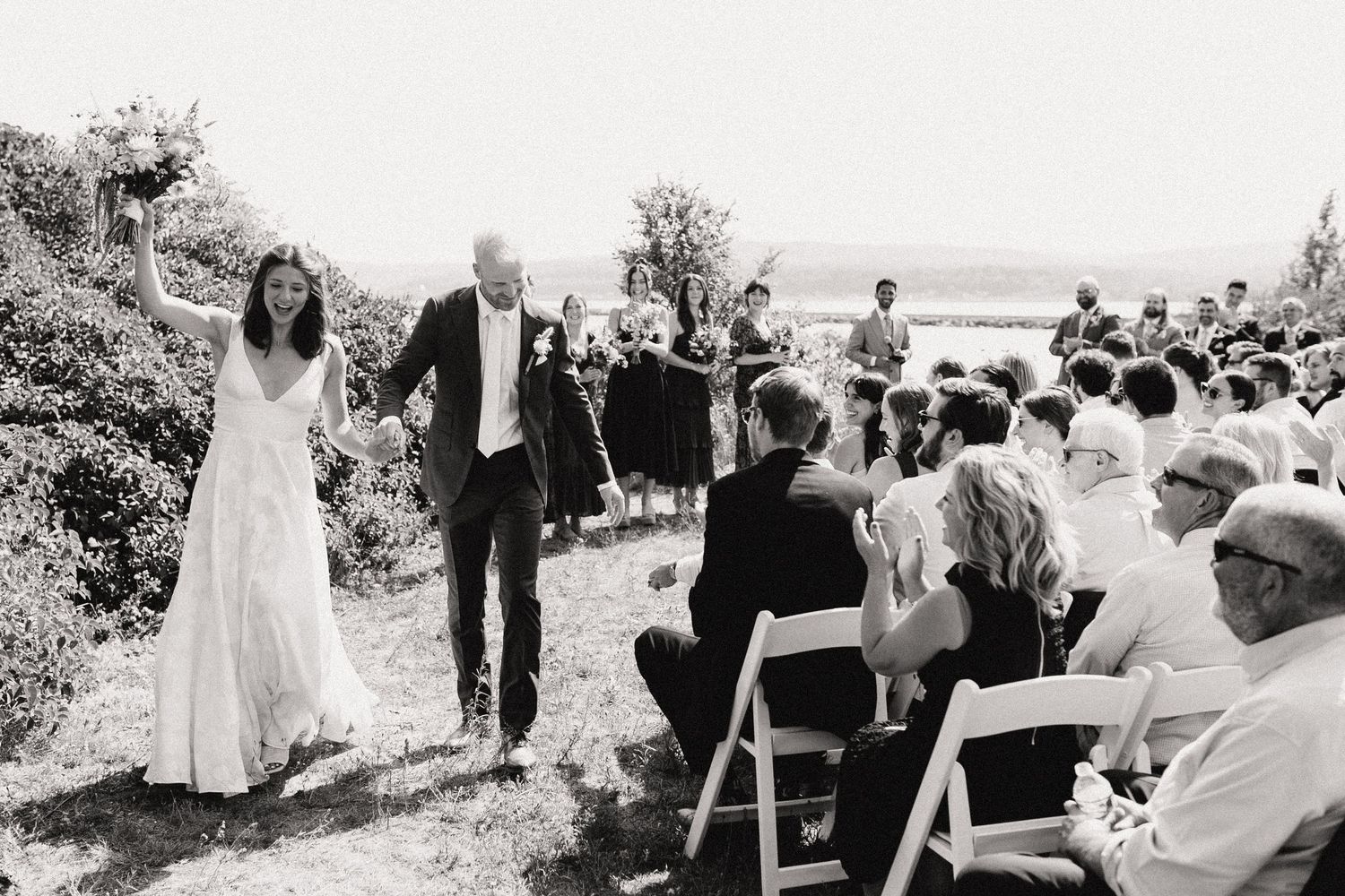 A bride and her father walk down the aisle at an outdoor wedding ceremony by the ocean.