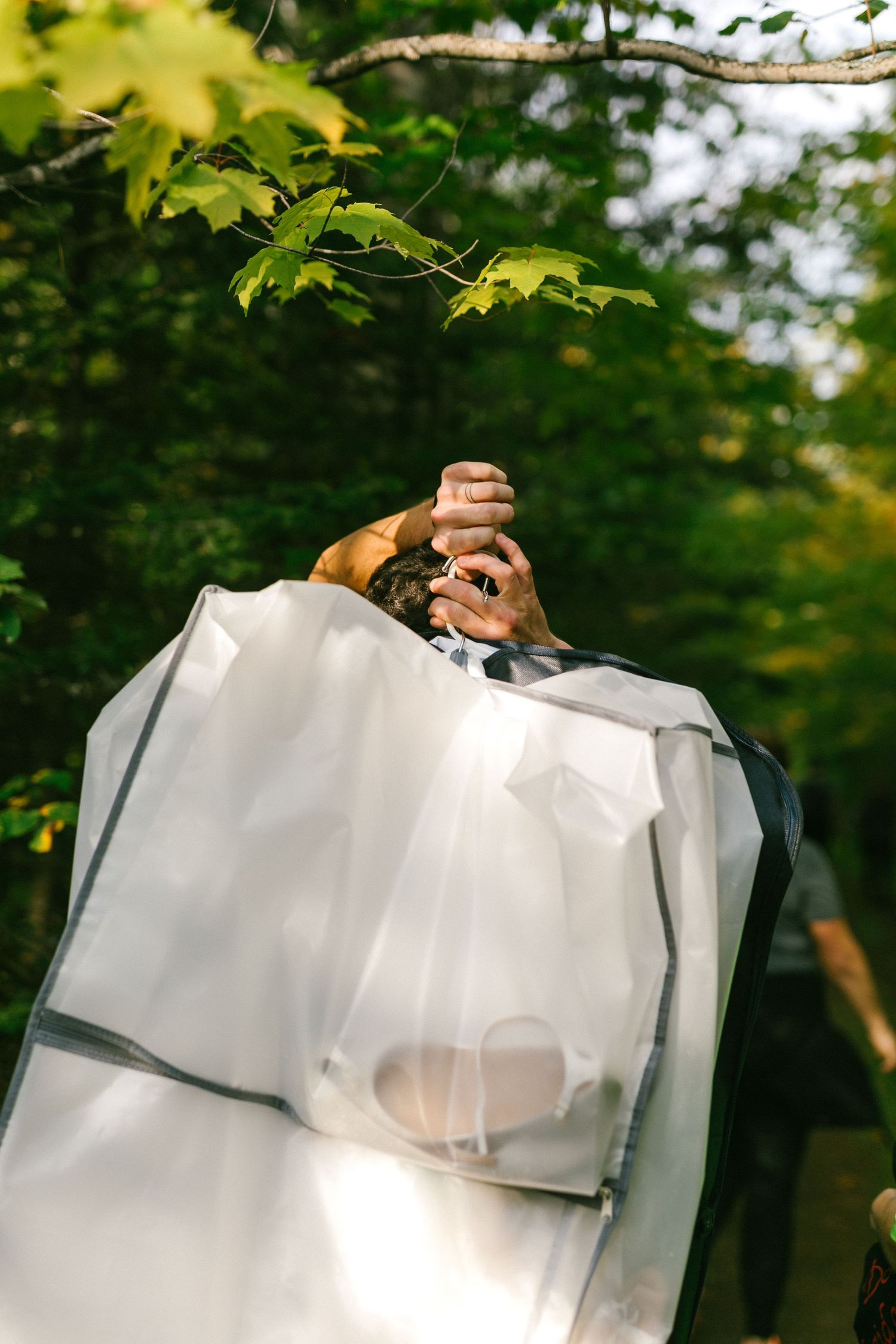 Close-up view of hands holding a garment bag with a dress visible through the translucent white material under maple leaves.