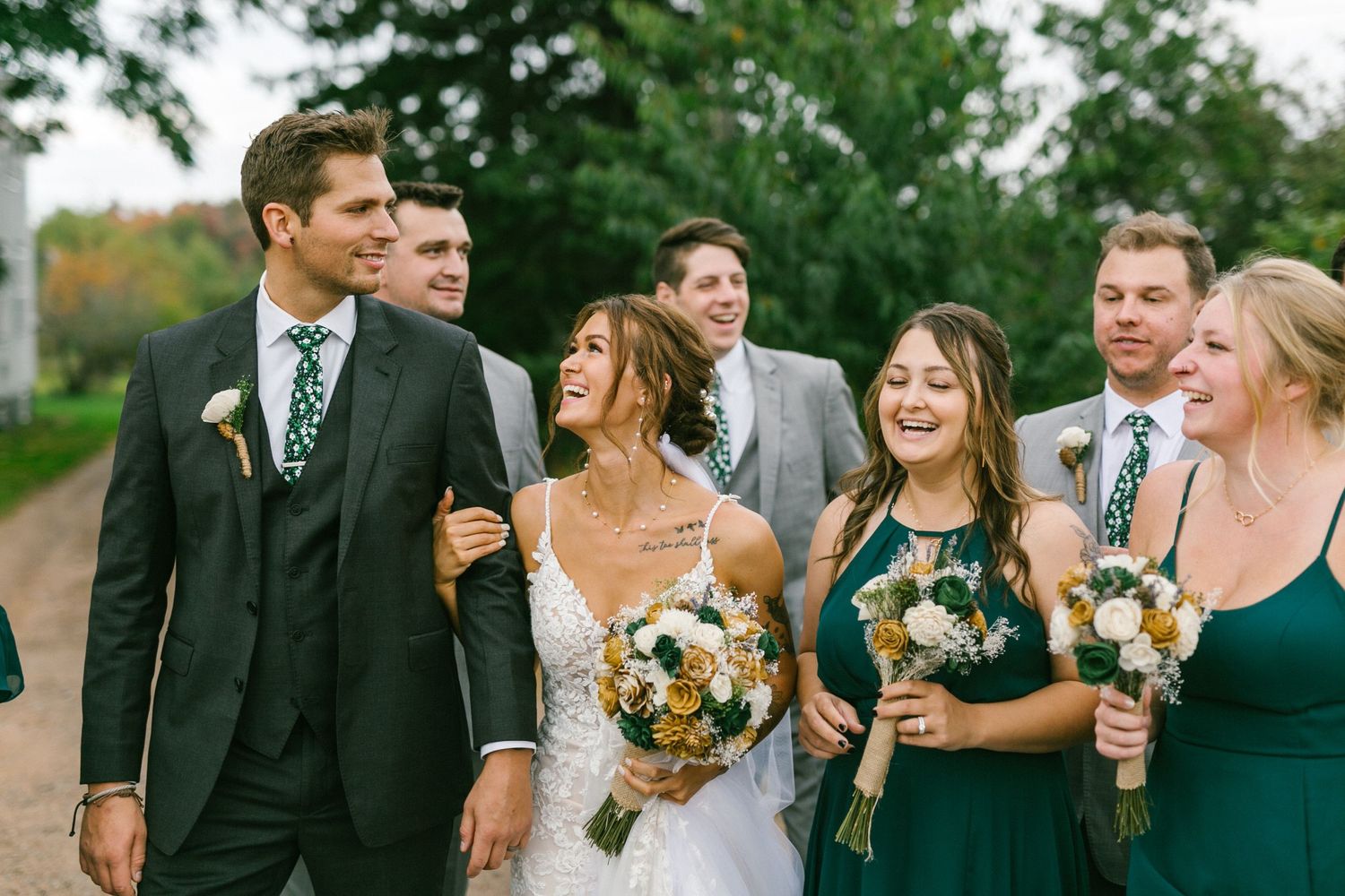 Wedding party walking and laughing together outdoors with bouquets along a tree-lined path.