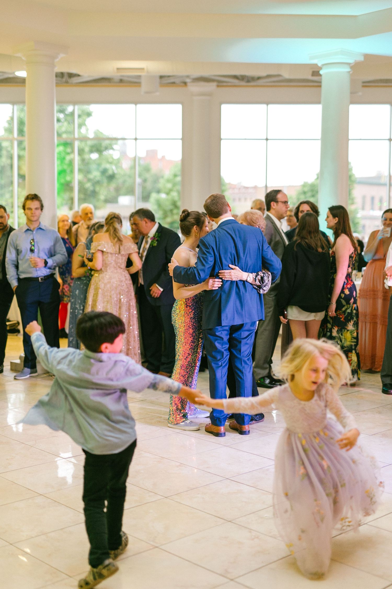 Young children dance together at a formal event while adults socialize in the background.