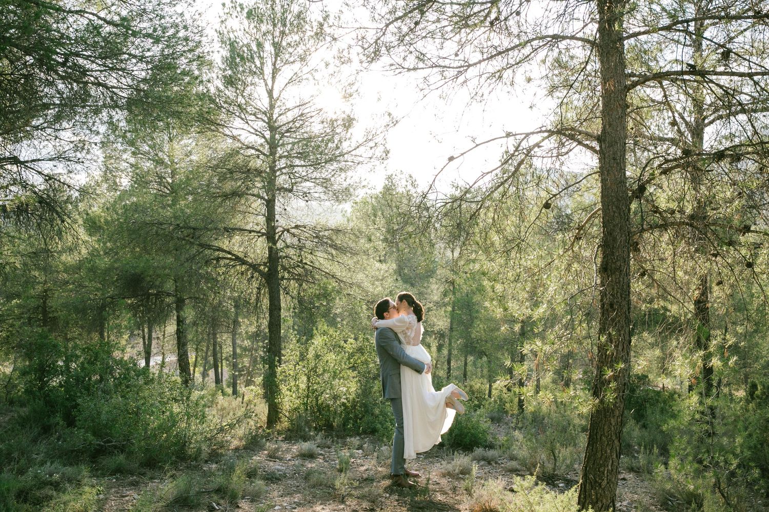 Couple embraces in serene sun-dappled forest wedding setting surrounded by tall pine trees. Spain destination wedding photographer.