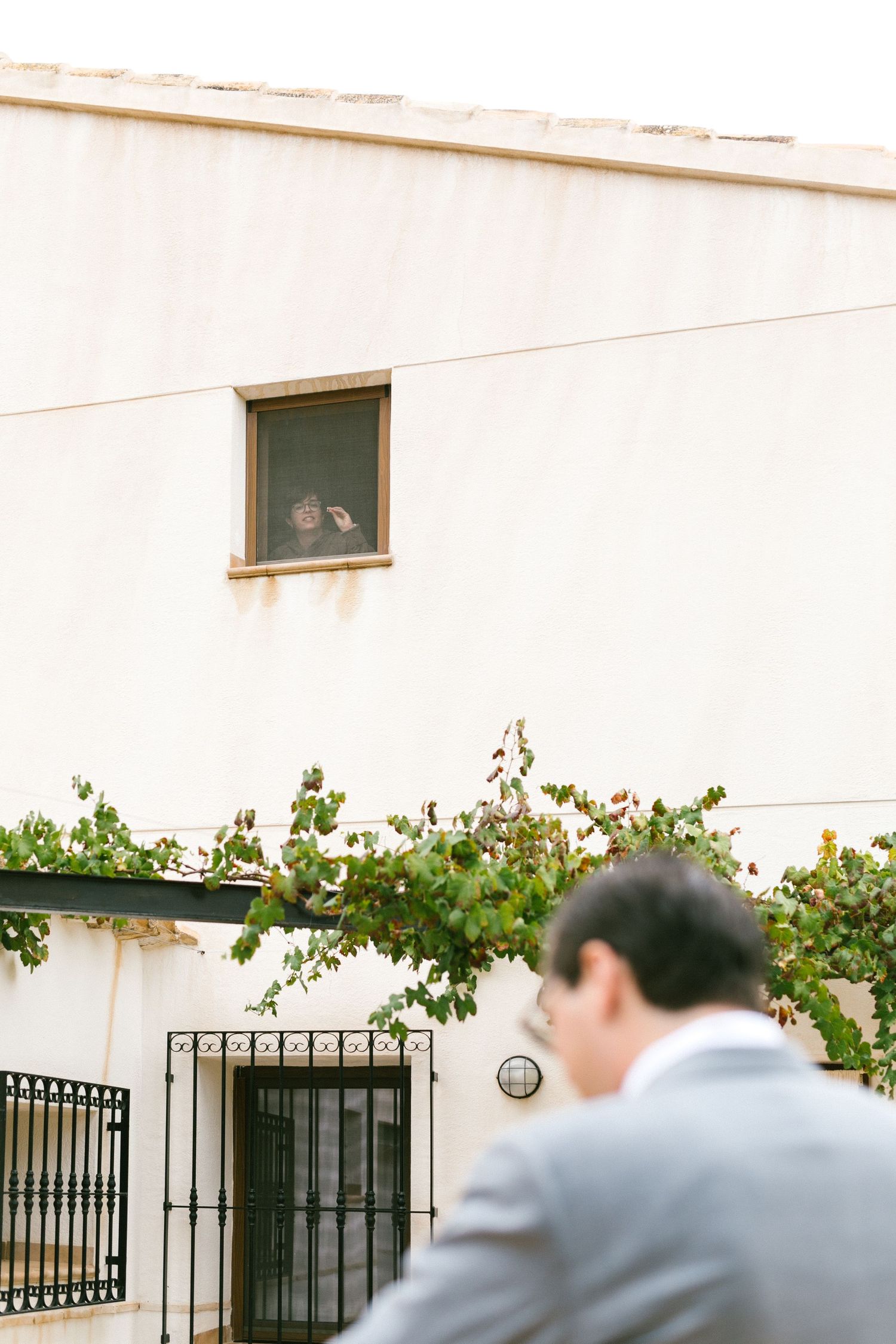 A grooms sister waves from a small window, near green climbing vines, and an iron gate entrance.