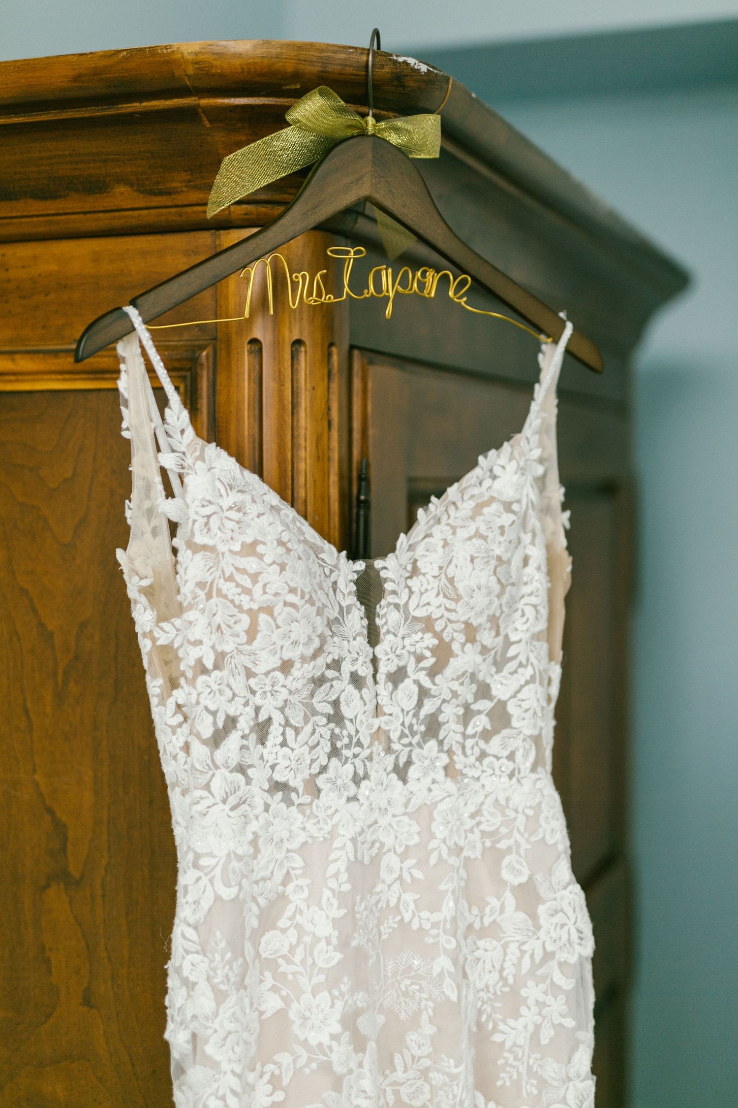 Elegant white lace wedding dress hanging on personalized hanger with green bow against wooden armoire.