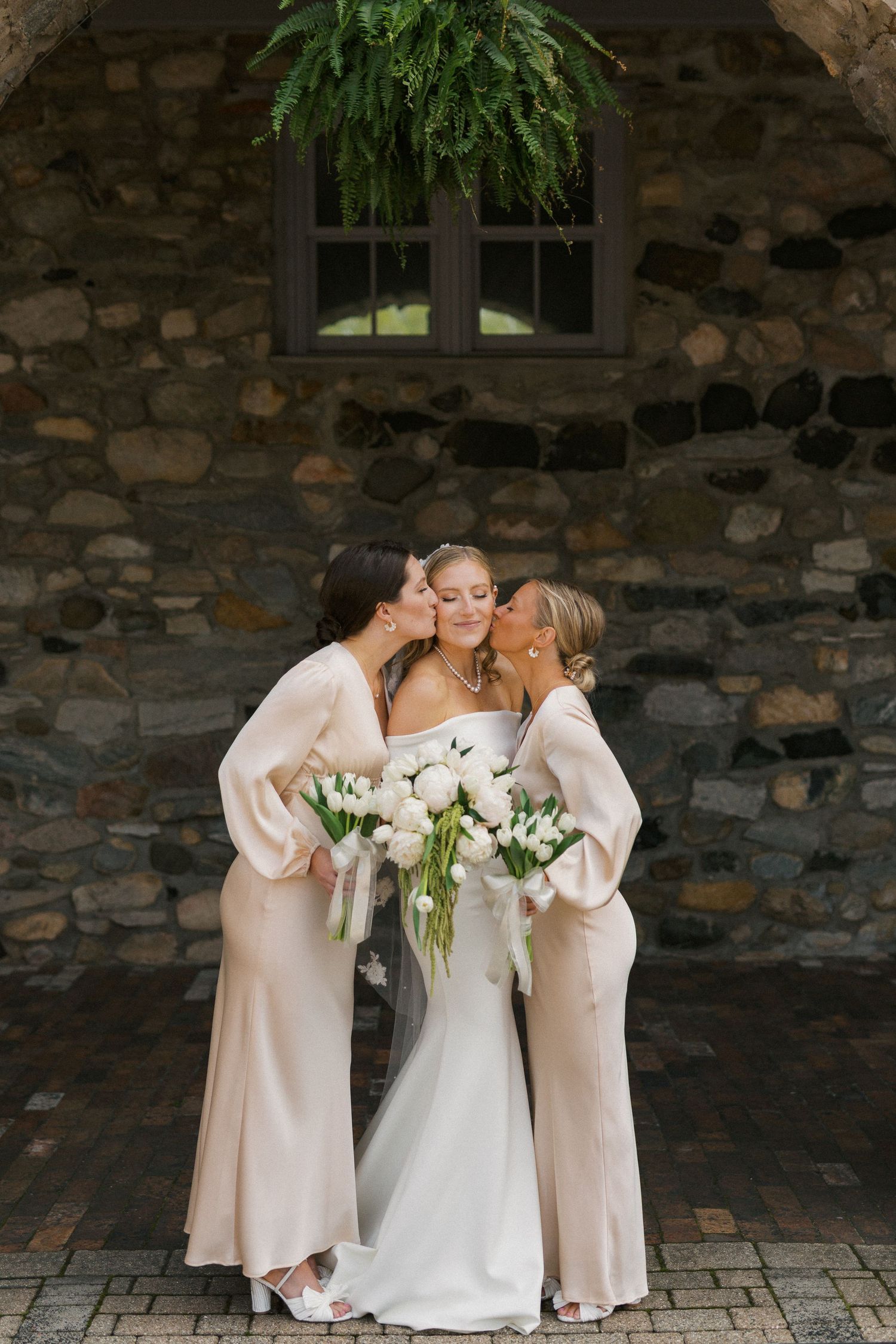 Bridesmaids in blush pink dresses sharing an intimate moment with bouquets against stone wall.