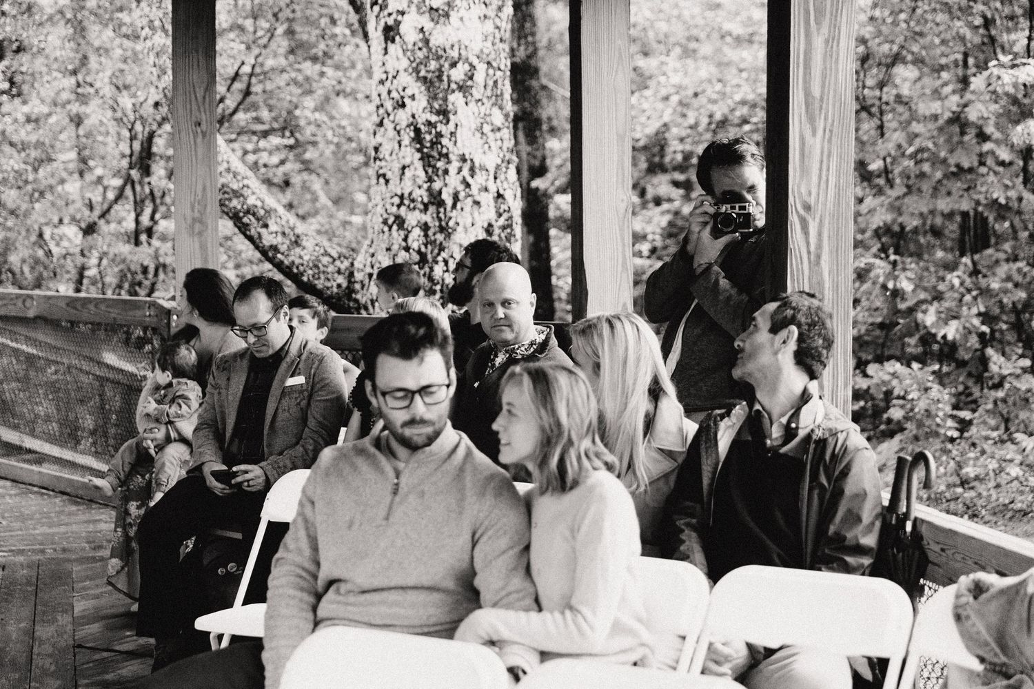 Black and white photo of wedding guests seated on wooden benches during outdoor ceremony.