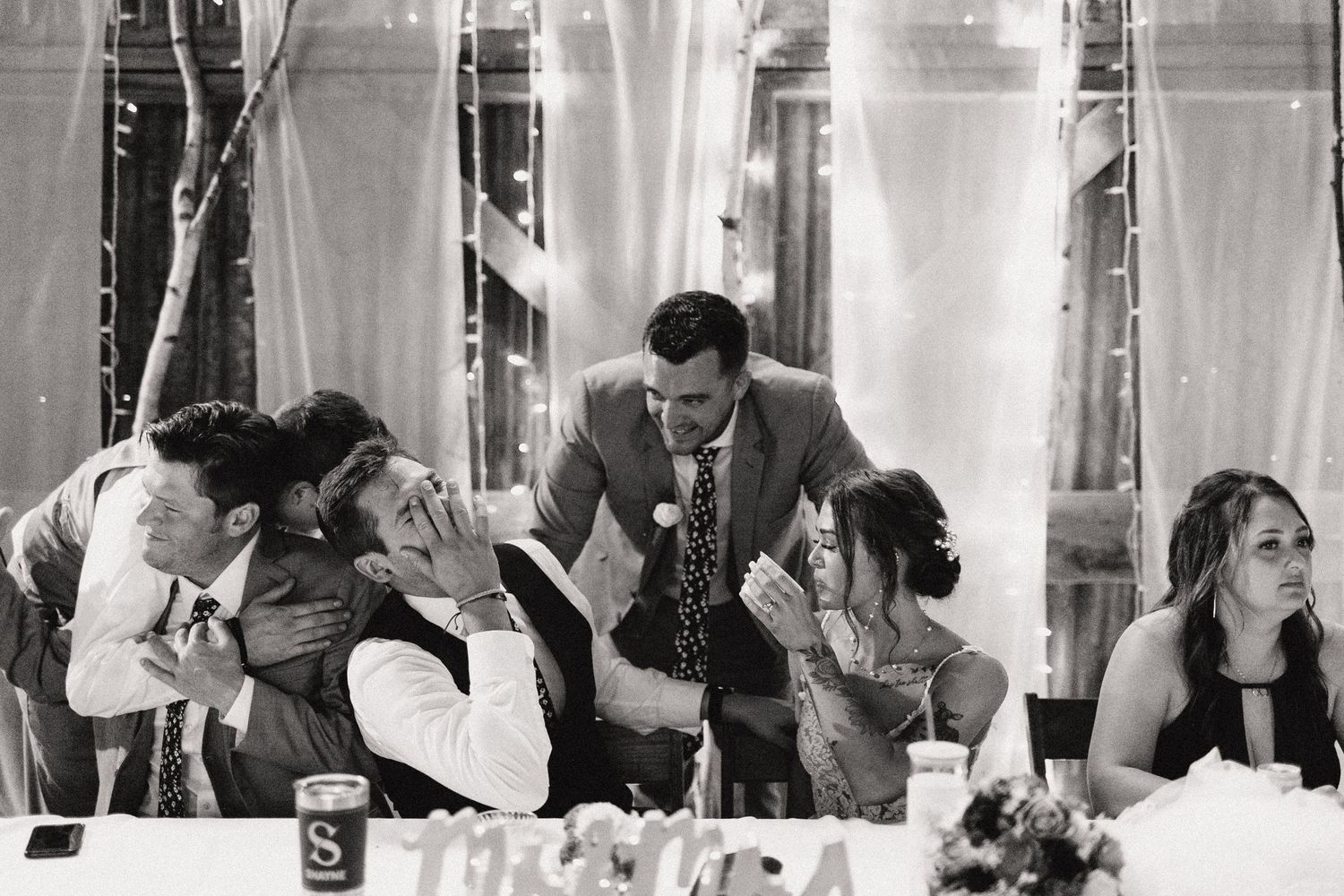 Black and white candid shot of wedding reception guests sharing laughs with bride and groom at decorated dinner tables.