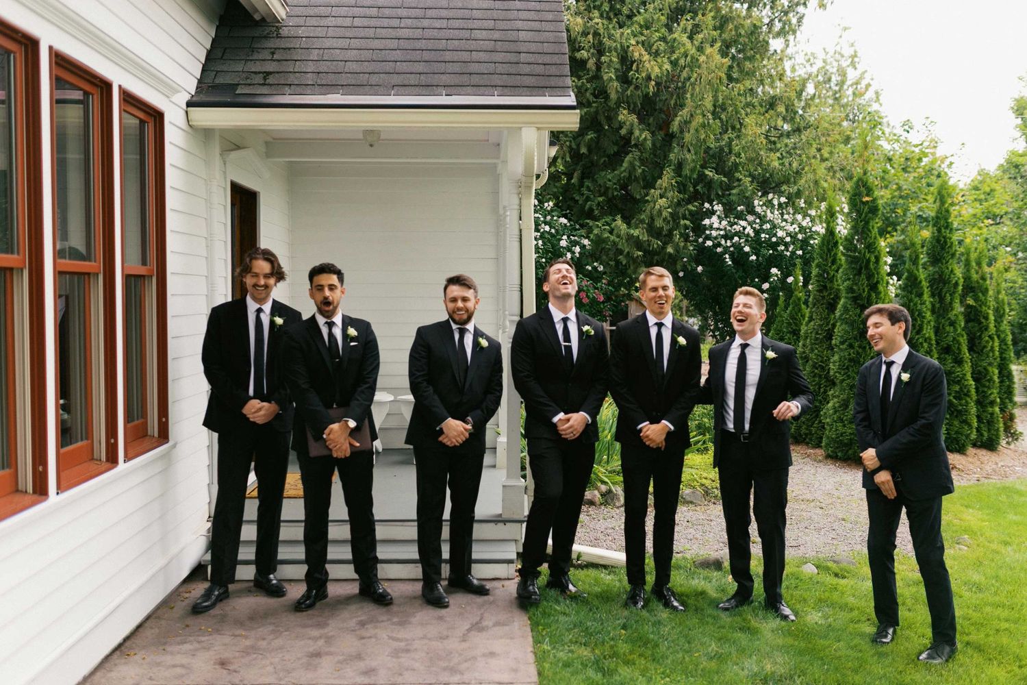 A wedding party in black suits stands in front of a white building with wooden trim on a rainy day.
