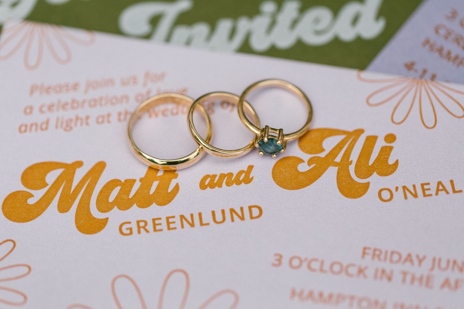 Three gold wedding rings arranged on a white and orange wedding invitation with text showing the couple's last name Greenlund.