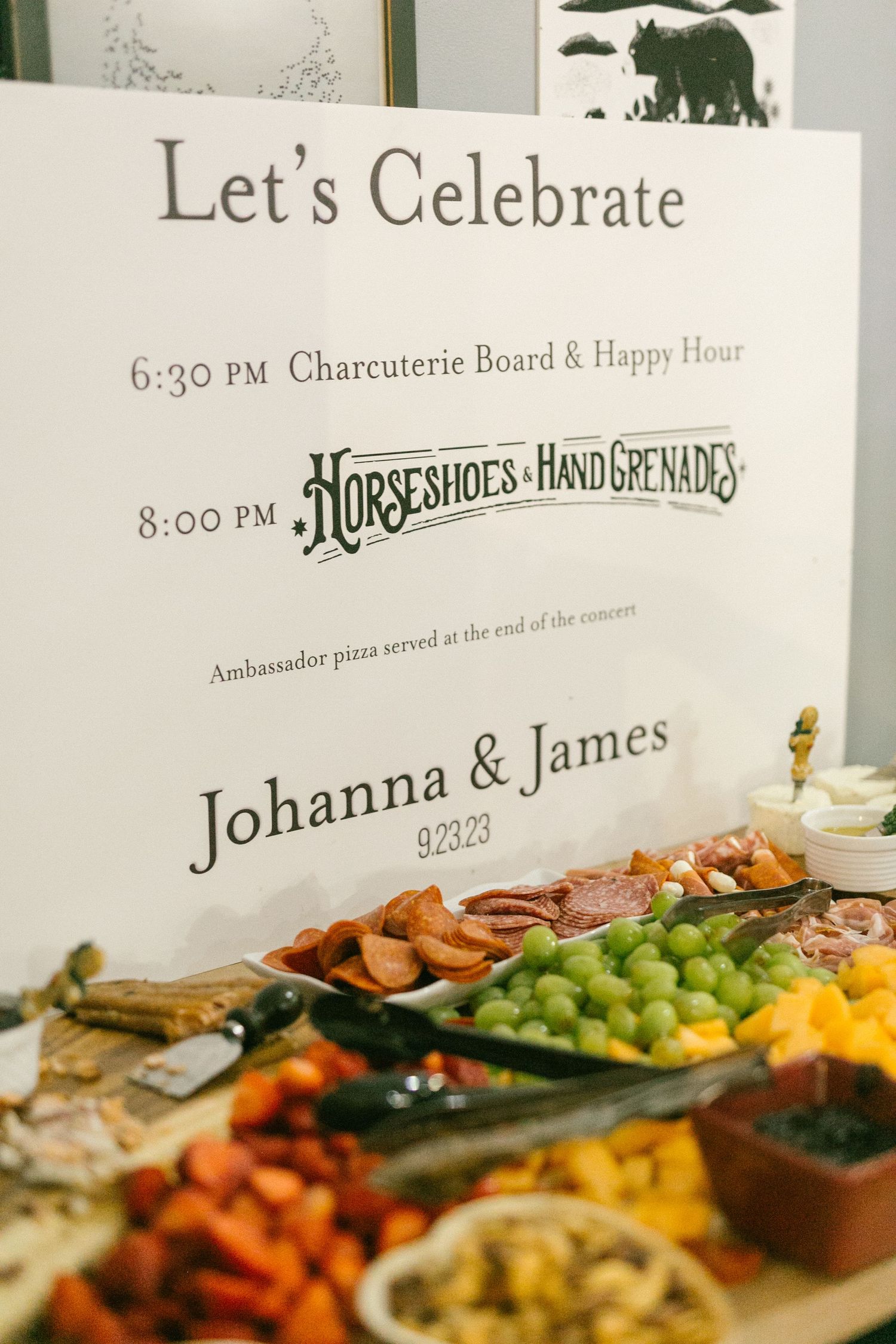 A wedding celebration sign displays timing details above an elegant charcuterie board with fresh fruits and appetizers.