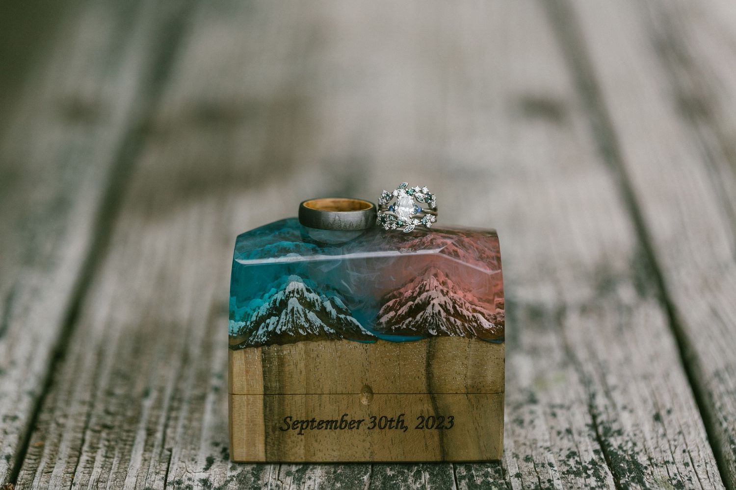 Wedding rings displayed on a wooden block with mountain scenery artwork and date engraving.