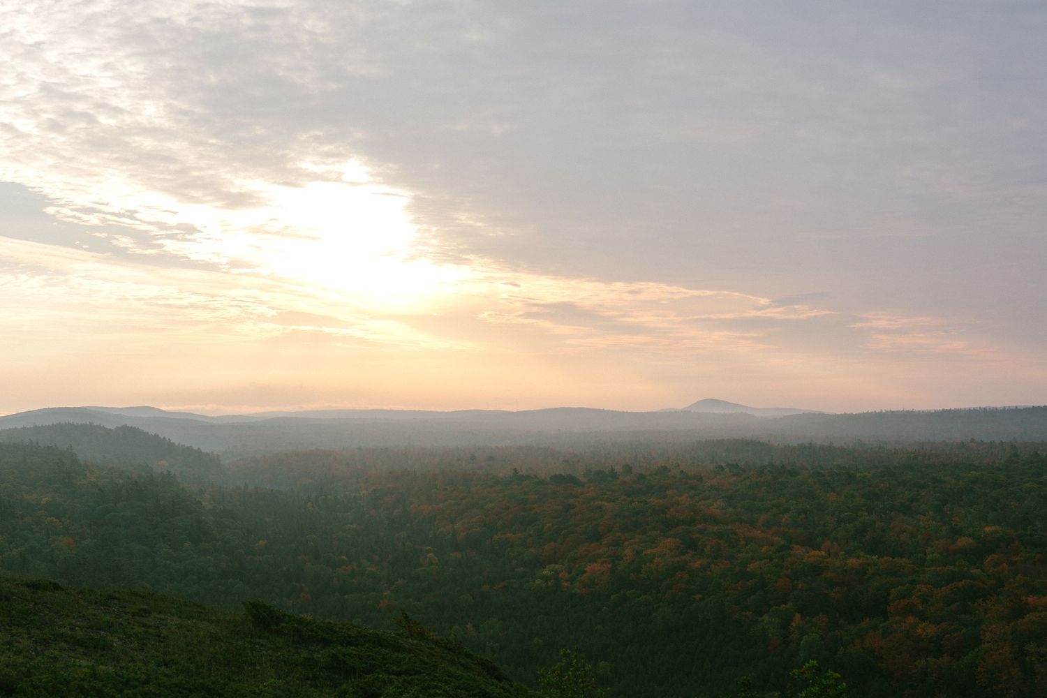 Misty mountain landscape at sunset with rolling hills and distant peaks silhouetted against a glowing evening sky.
