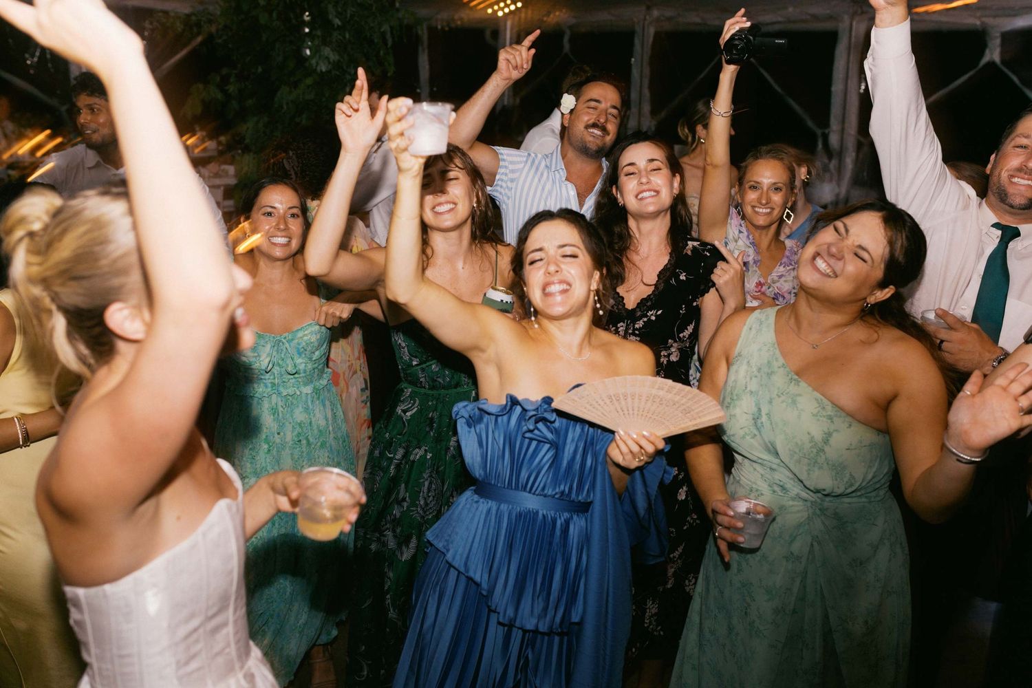 Wedding guests dancing and celebrating with arms raised next to a joyous bride on a dimly lit dance floor at a reception party.