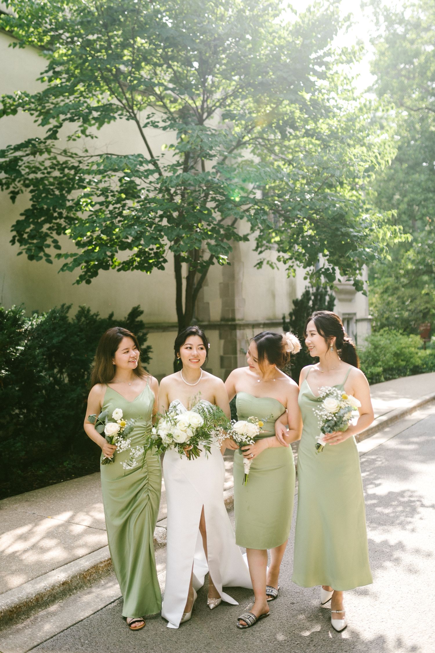 Bride and three bridesmaids in sage green dresses holding white bouquets walking outdoors on wedding day.