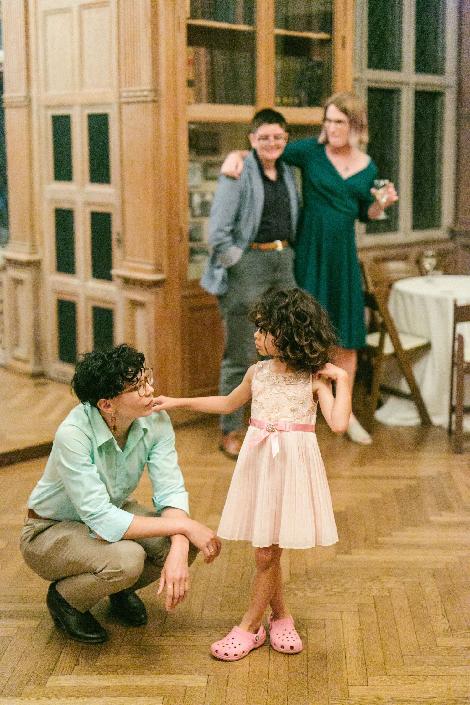 Adult crouching to interact with young girl in pink dress and crocs at elegant wedding reception venue.