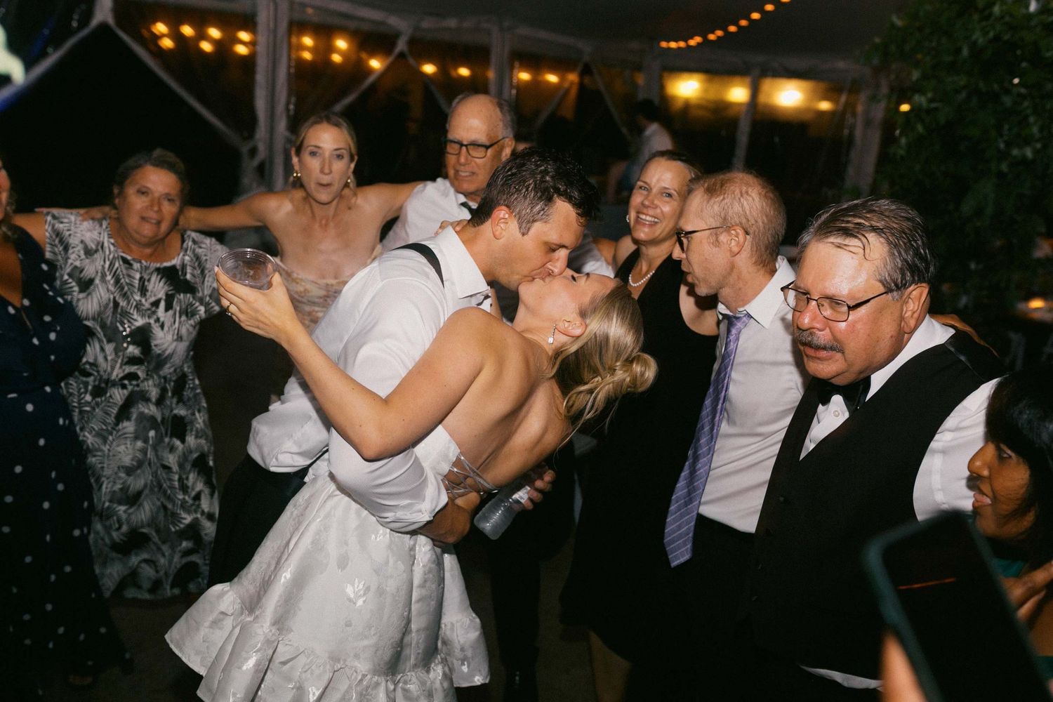 Couple shares romantic kiss on dance floor at evening wedding reception while guests look on with smiles and delight.
