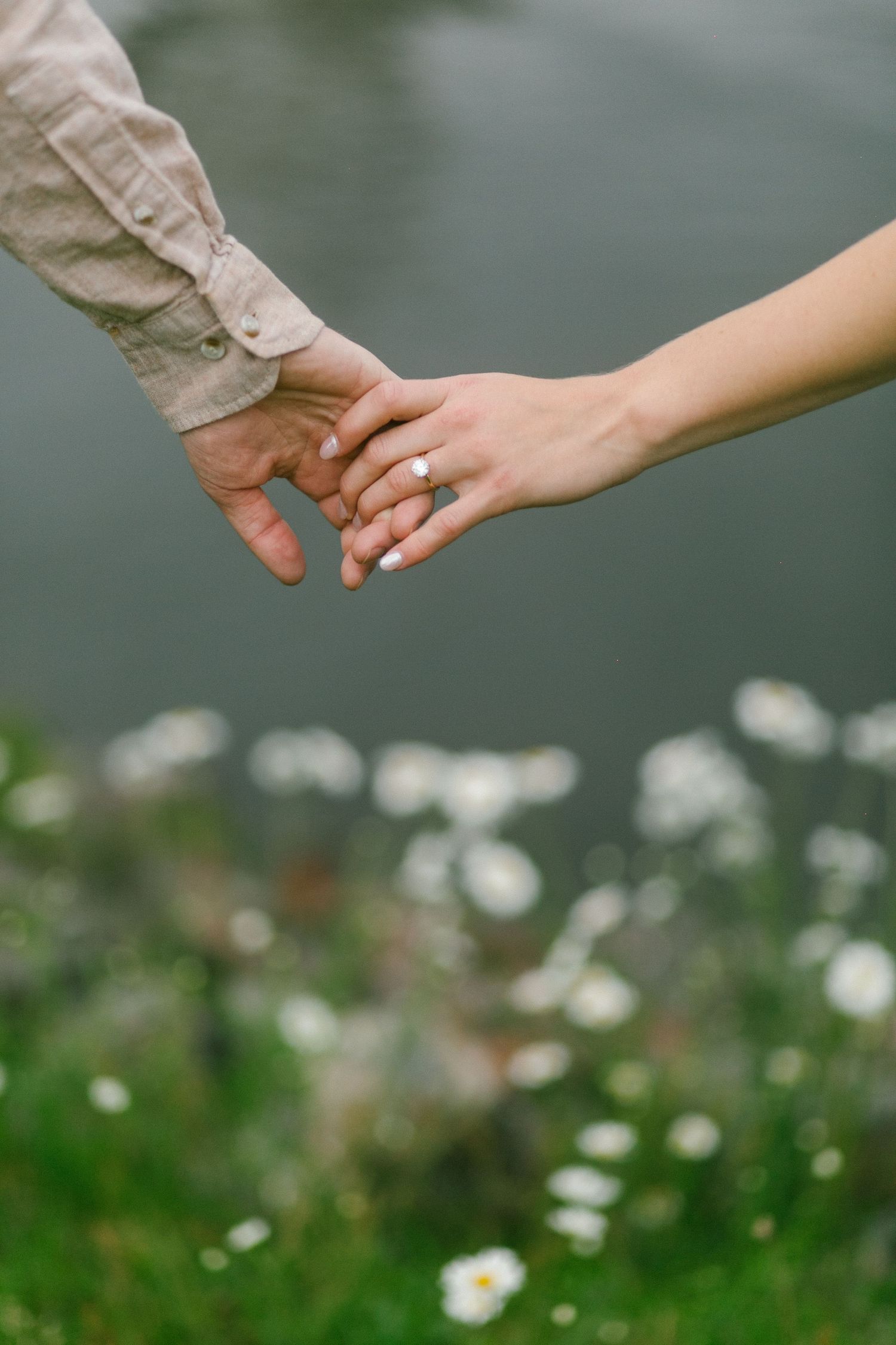 Two hands gently touch with a sparkling engagement ring against a backdrop of white wildflowers and blurred water.