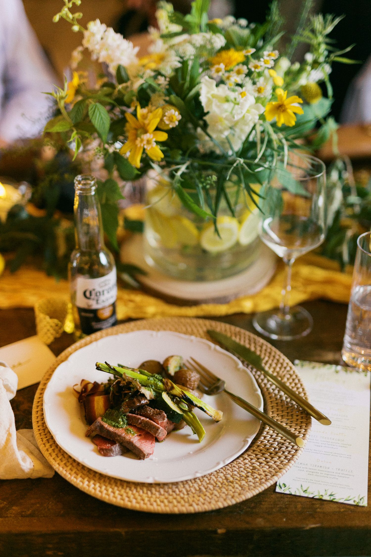Elegant wedding reception place setting with sliced steak, asparagus, Corona beer, and yellow wildflower centerpiece on rustic wood table.
