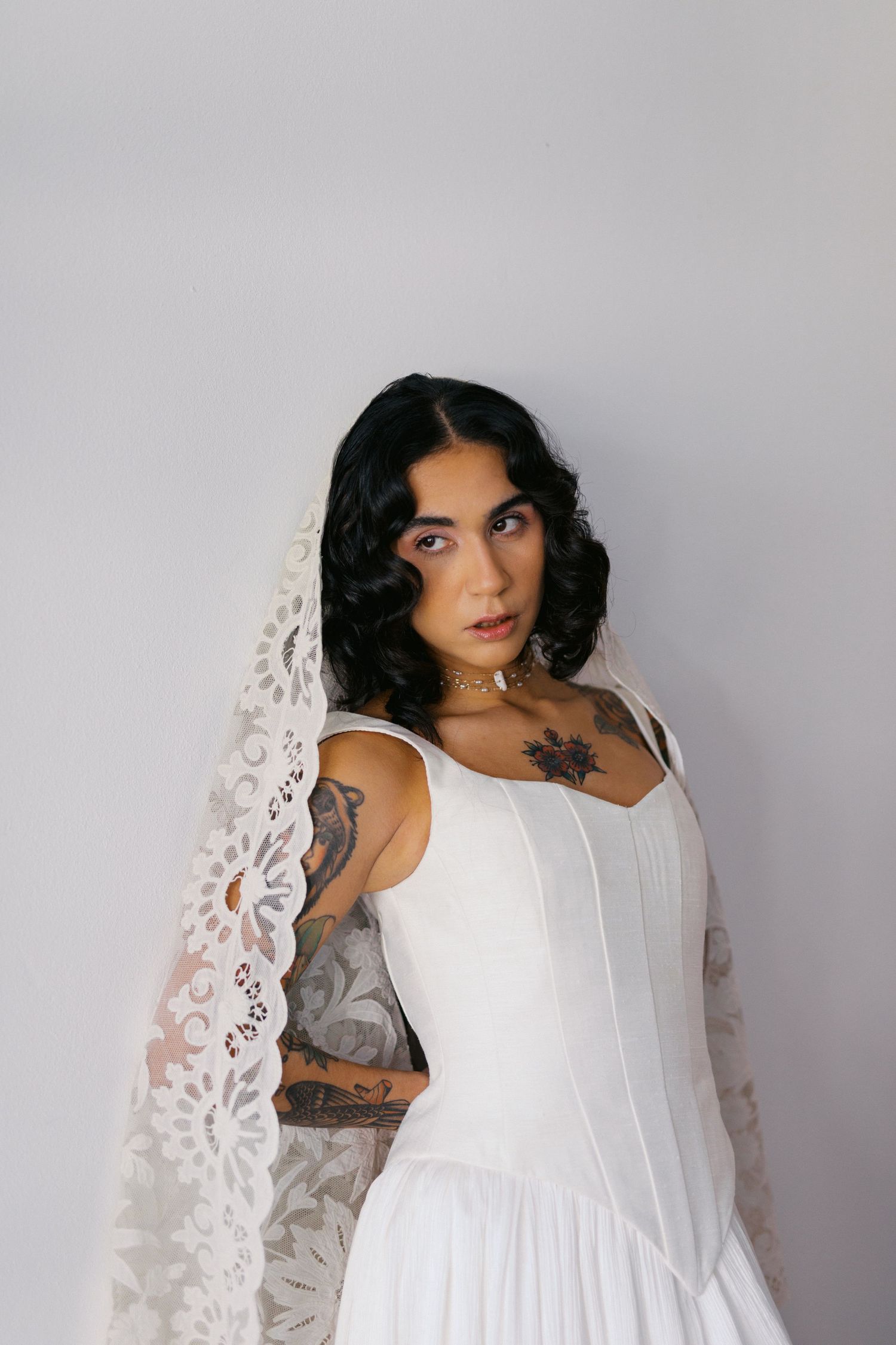 Tattooed bride in white corset gown with intricate lace veil, dark curly hair, and gold choker necklace.
