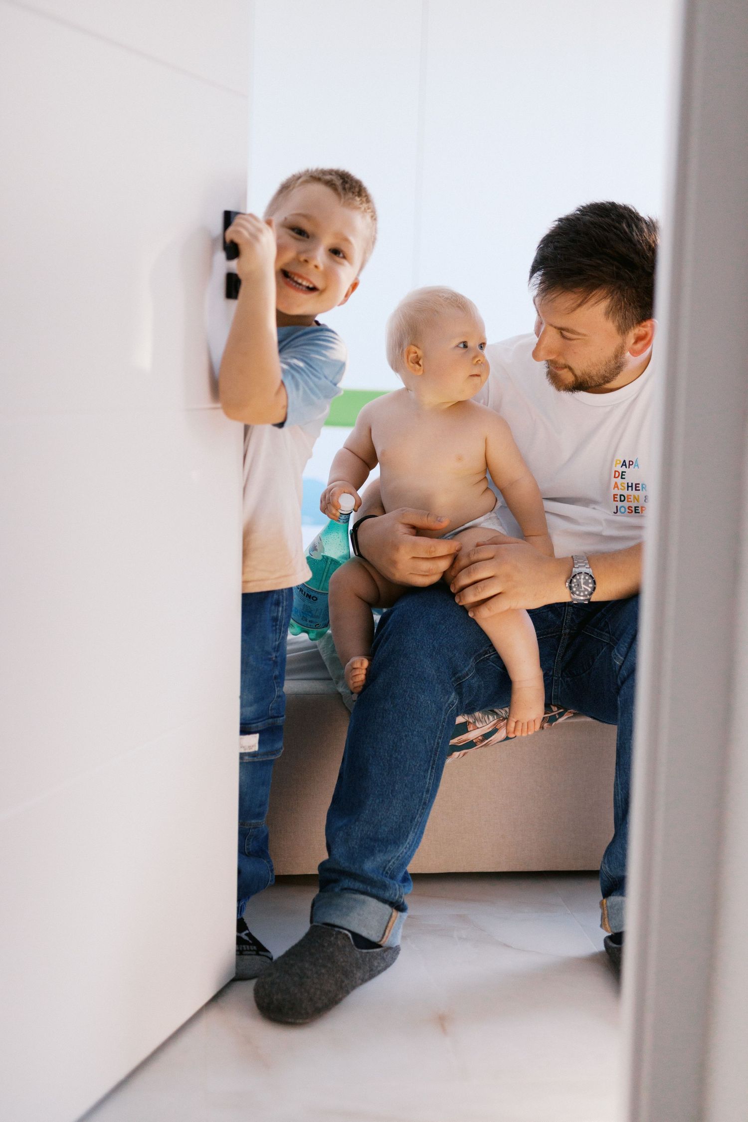 A father sits with his baby on his lap while an older boy plays nearby in a bright white room.