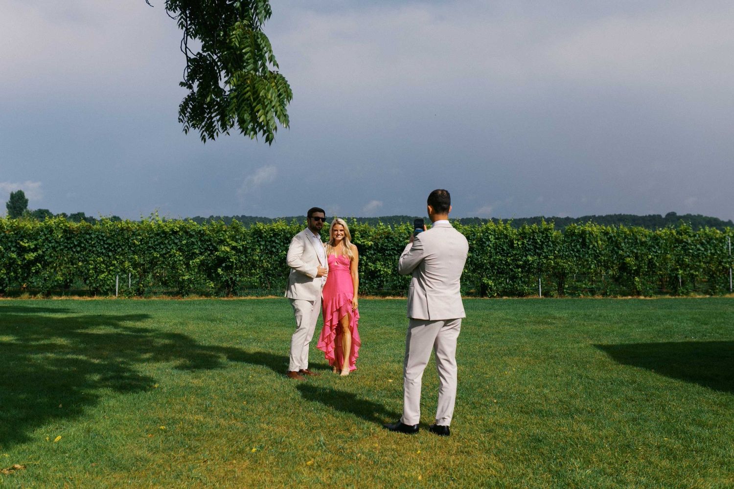 People in wedding attire stand for a photo on a lush green lawn with sunflower fields stretching into the horizon.