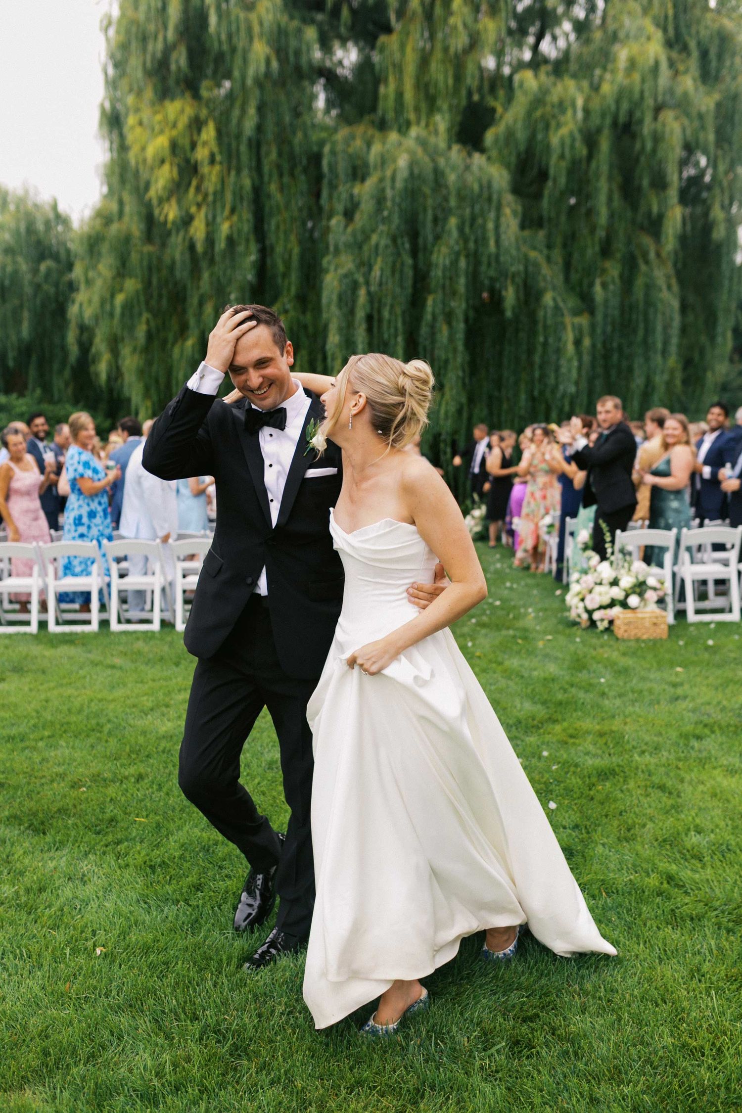 A couple in wedding attire dances playfully on a green lawn with weeping willow trees and guests in the background.