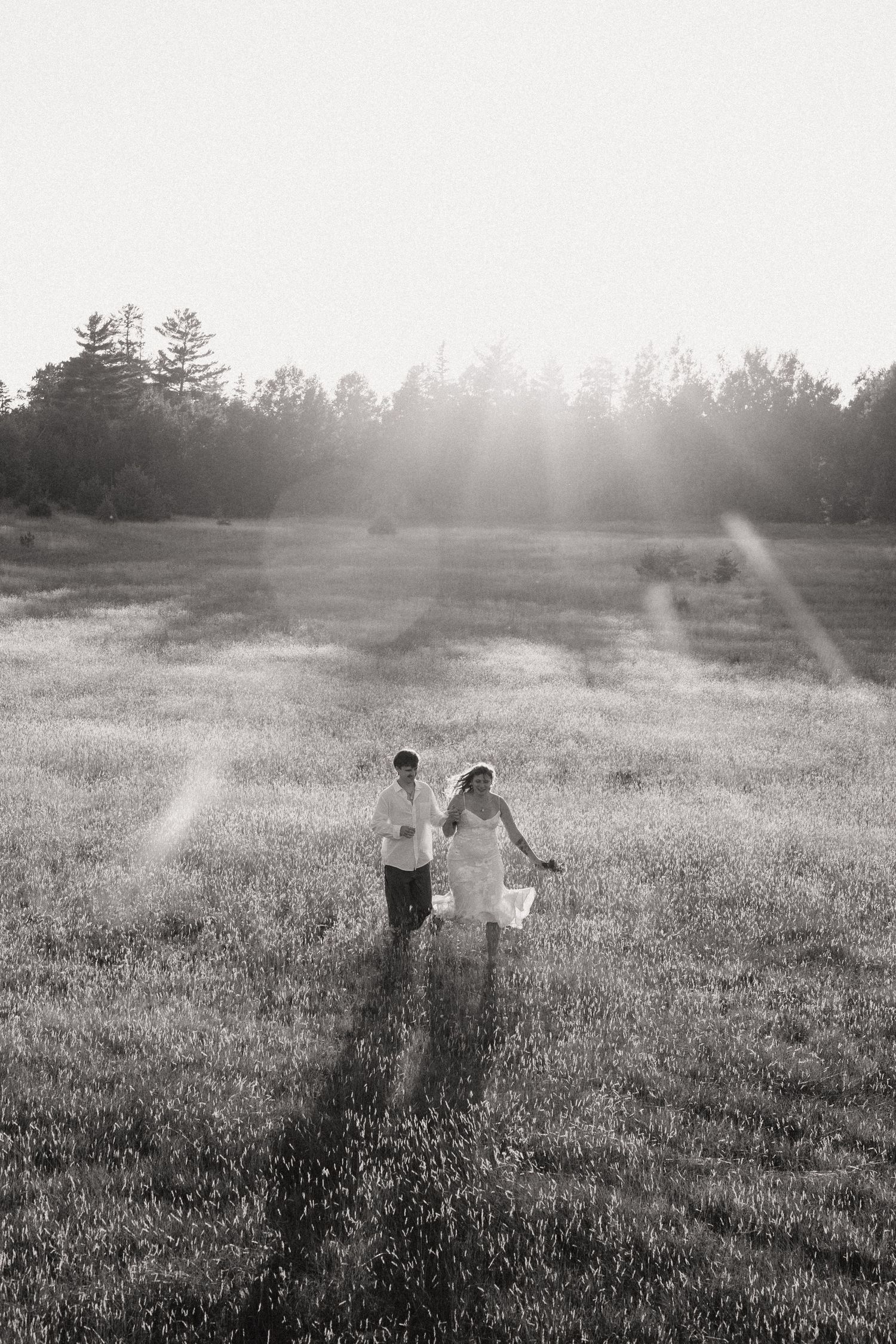 Black and white photo of a bride and groom holding hands in a sunlit field with trees in the background.