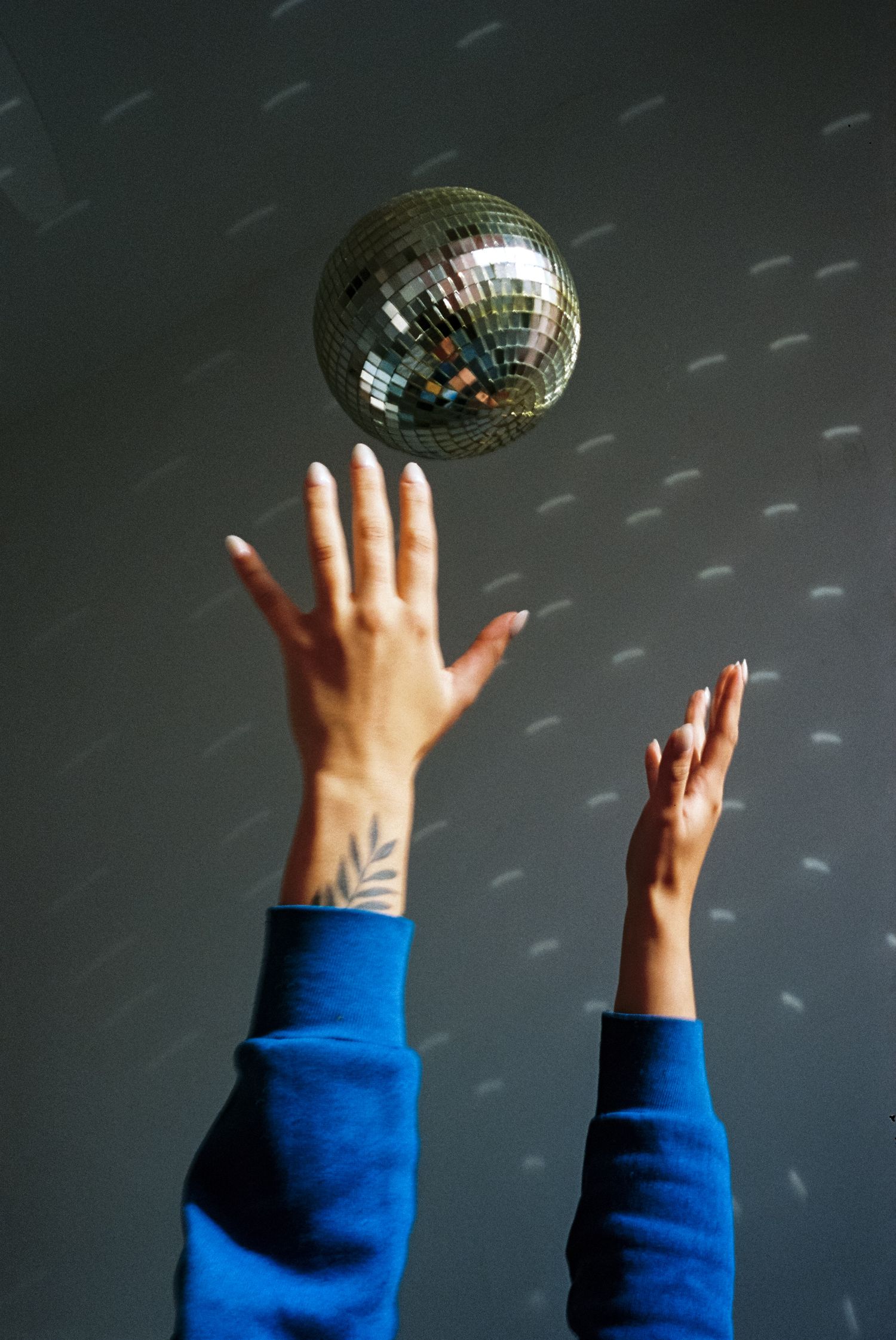 Hands reaching up toward a sparkling disco ball against a gray wall in blue sleeves.