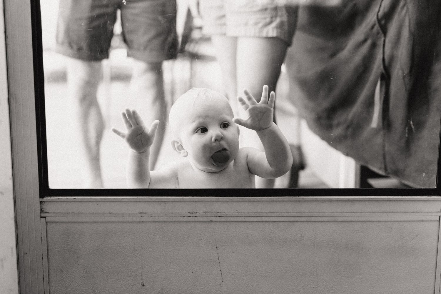 Curious baby pressing hands and face against a glass door in black and white photo.