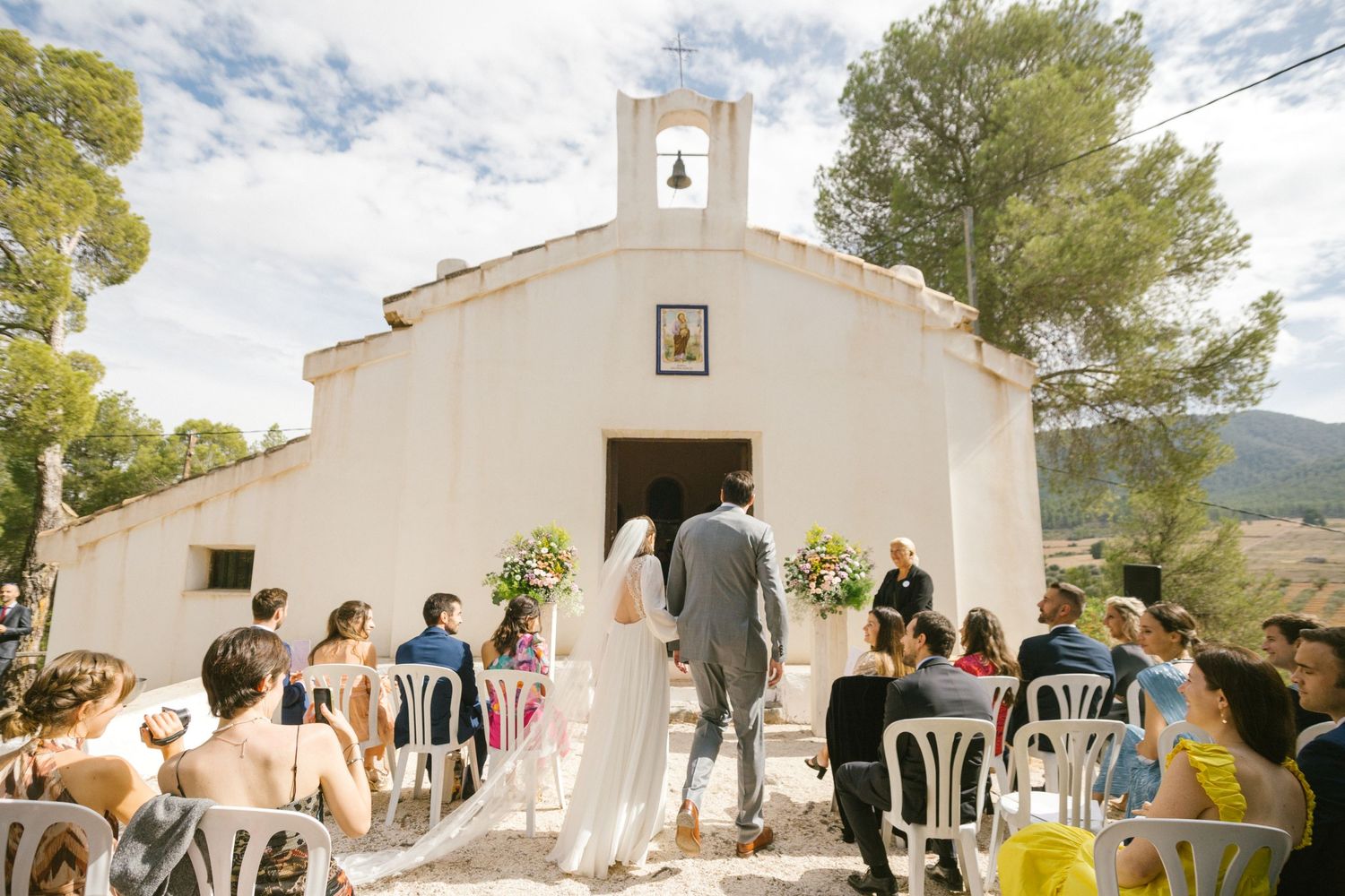 Bride and groom walk into a quaint white chapel with mountains in the distance in Murcia Spain.