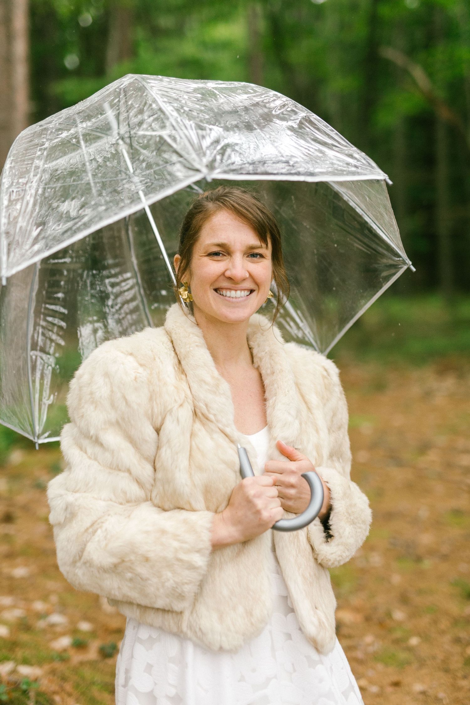 A person in a white dress and cream fur jacket holds a clear umbrella while standing in autumn woods.