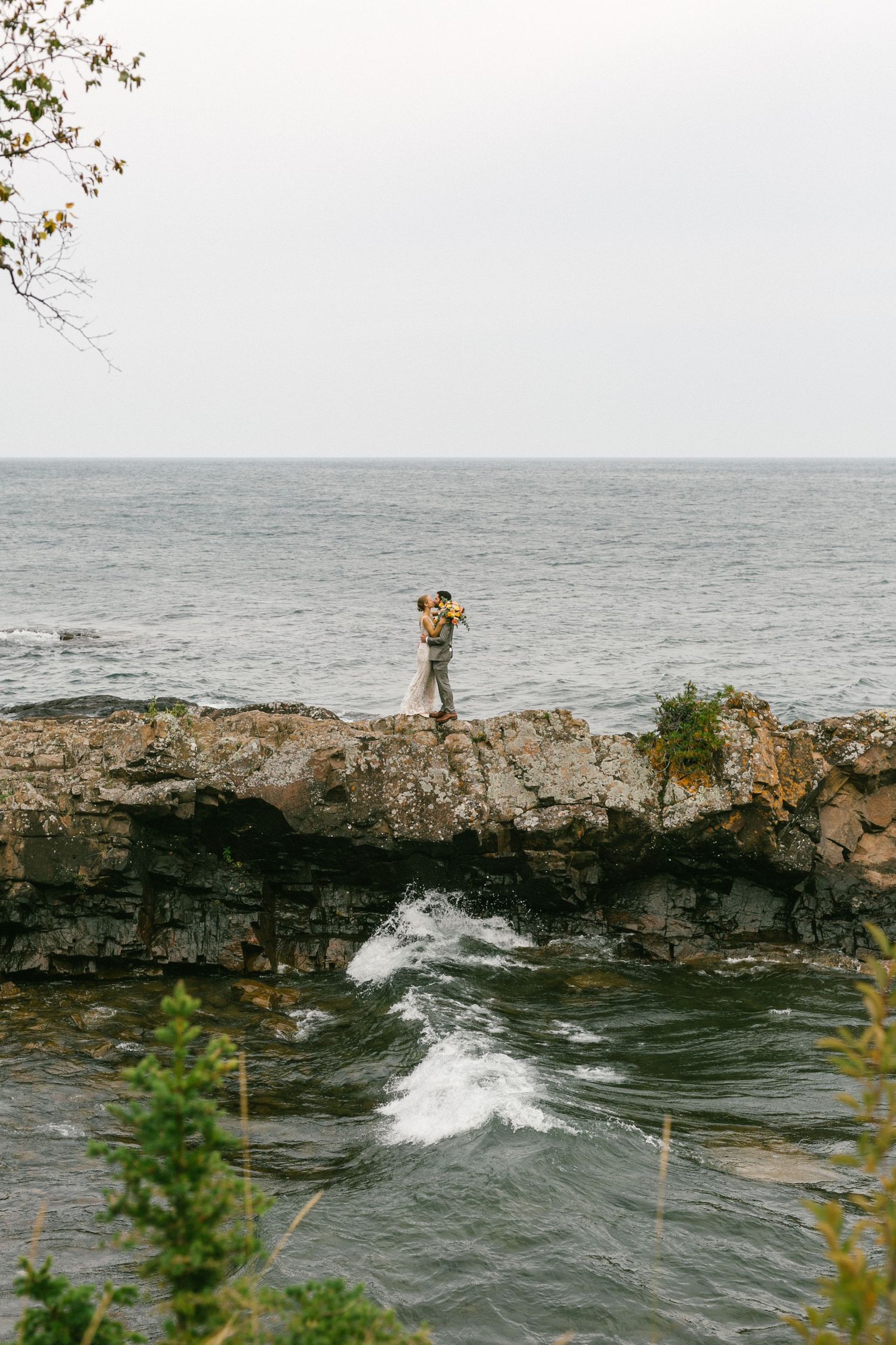 Someone stands on rugged coastal cliffs overlooking turbulent ocean waves breaking against rocky shoreline on an overcast day.