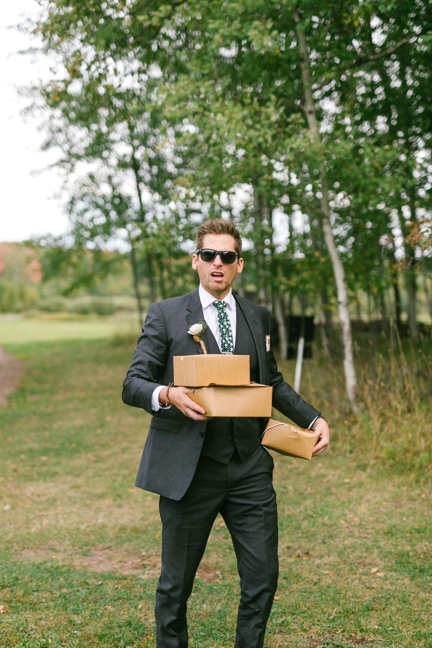 Well-dressed groom in dark suit and sunglasses carrying brown cardboard boxes outdoors near trees in a grassy area.