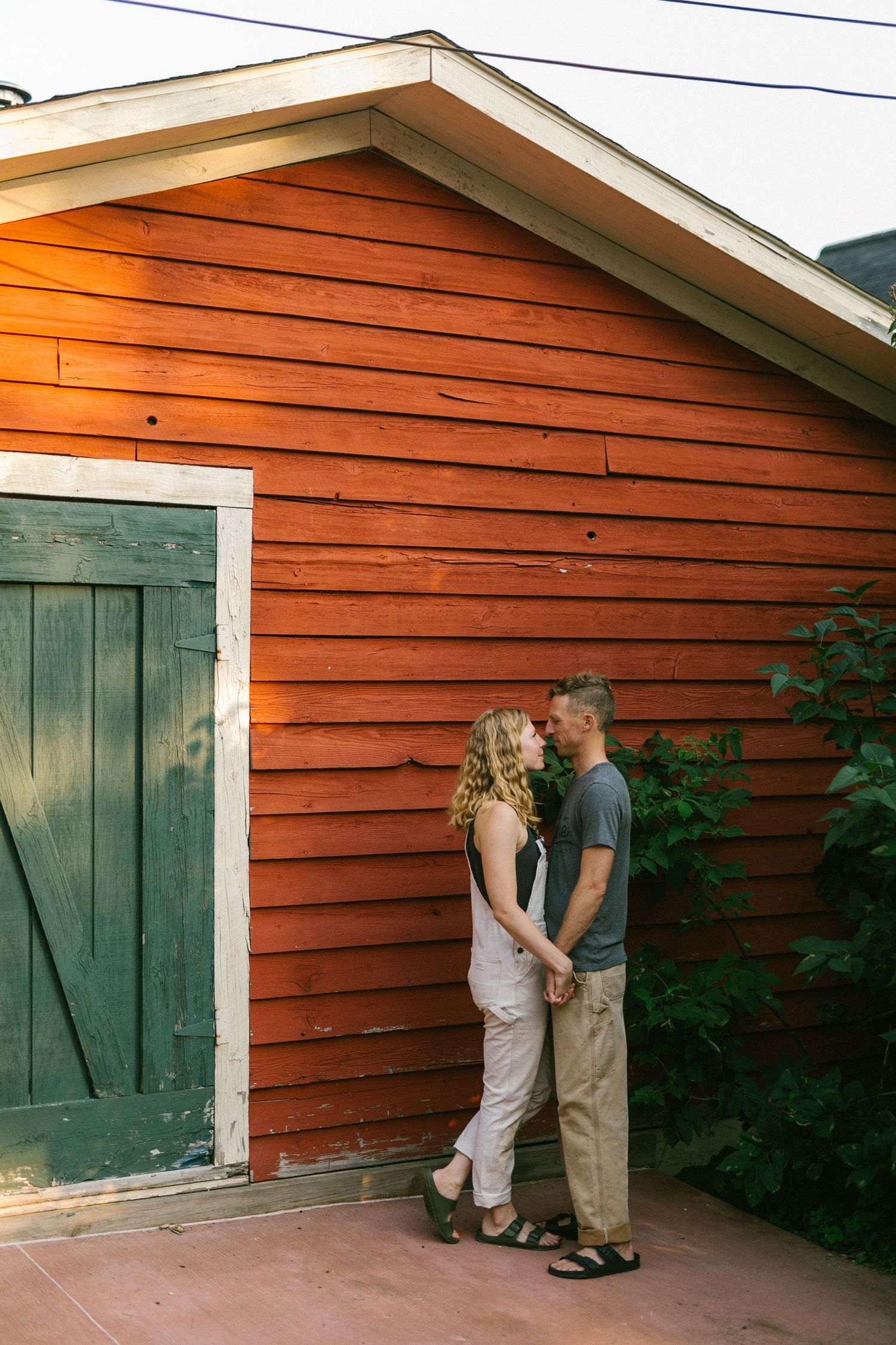 Couple stands intimately against rustic orange siding near a green barn door.