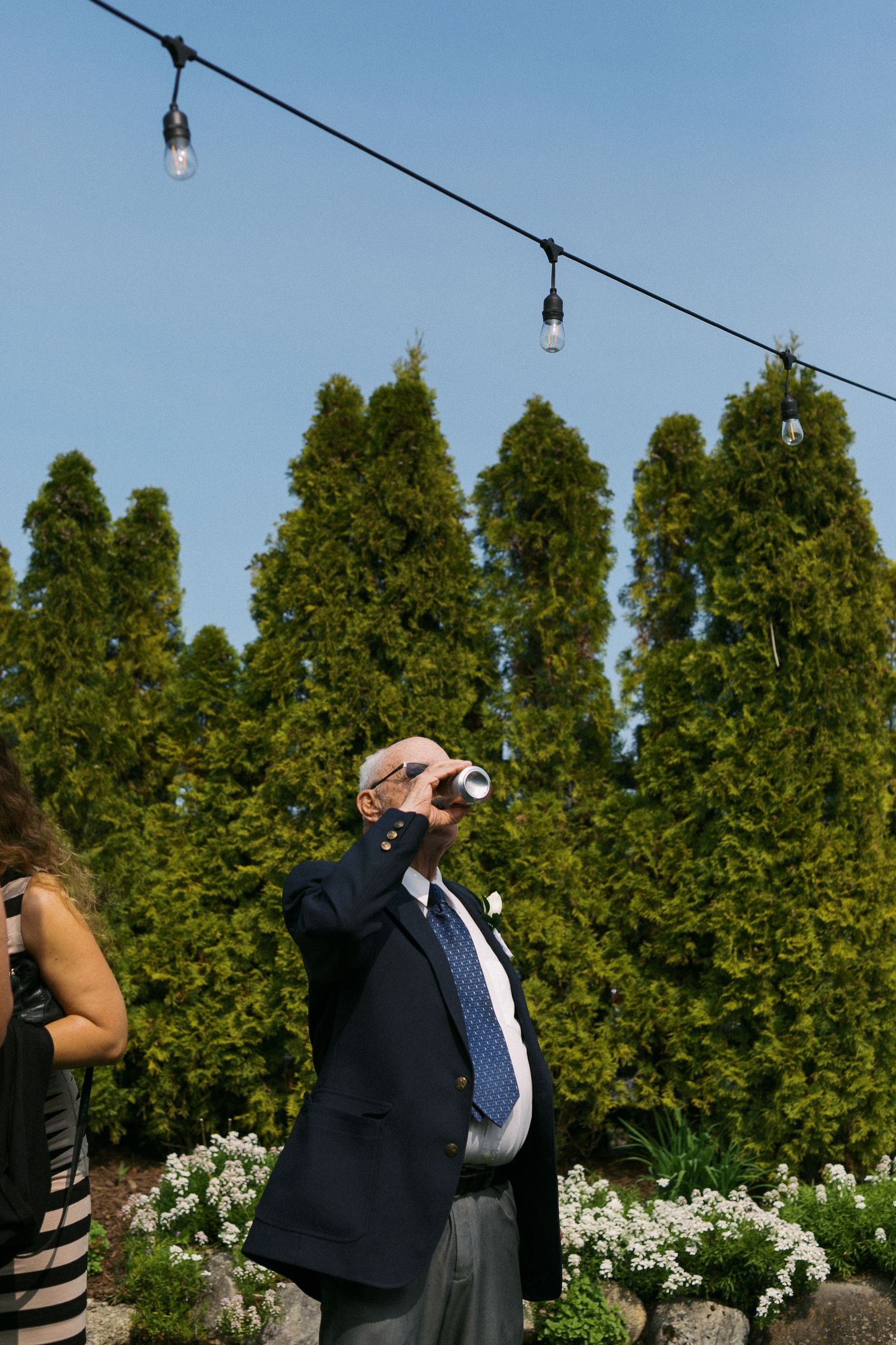 A guest at an outdoor event drinks from a glass under string lights with evergreen trees in the background.