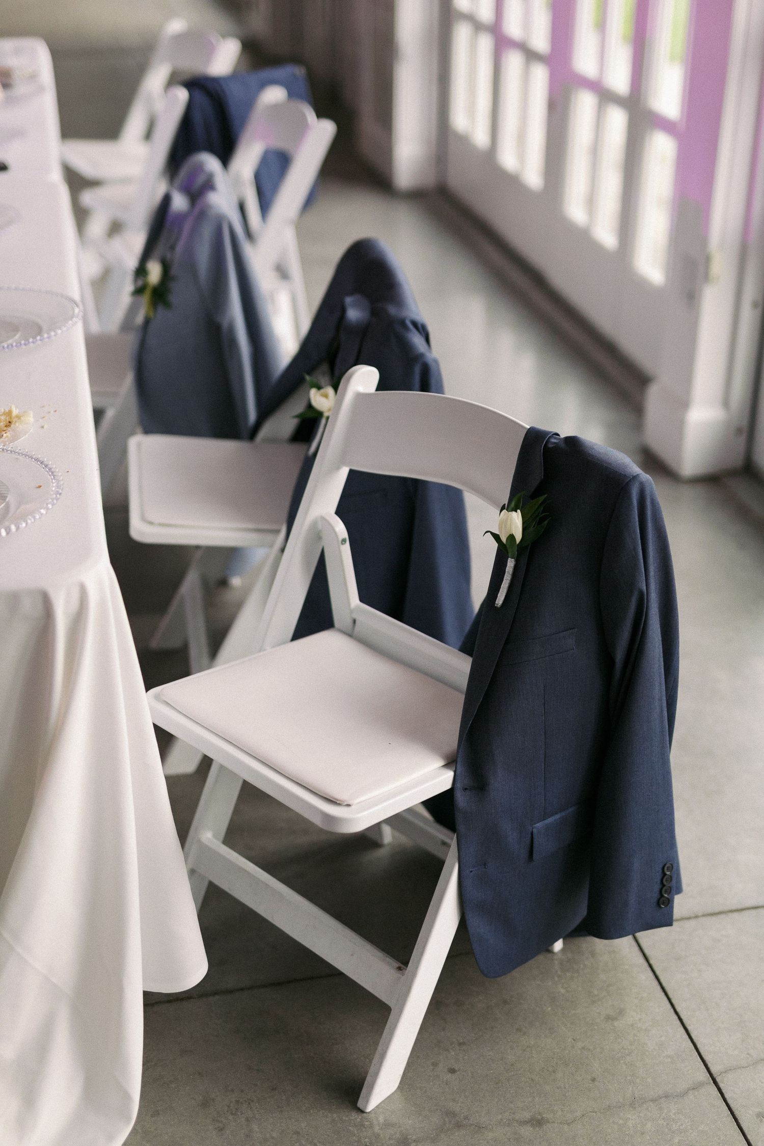 White folding chairs and navy suit jackets arranged for a wedding ceremony indoors.