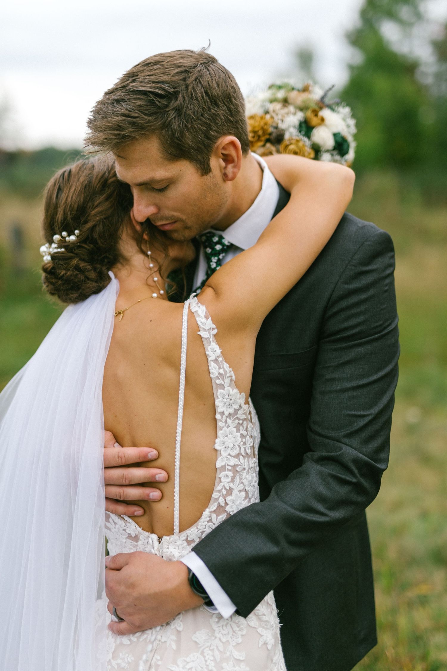 Bride in backless lace gown embraces groom during intimate outdoor wedding photoshoot.