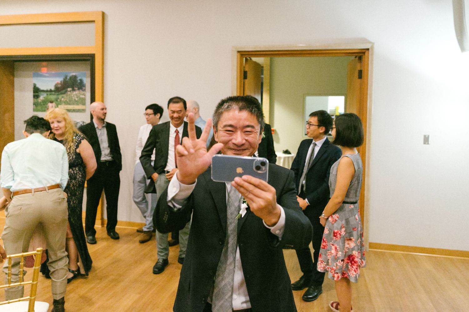 Man in suit taking selfie with peace sign gesture at indoor social gathering with guests mingling in background.