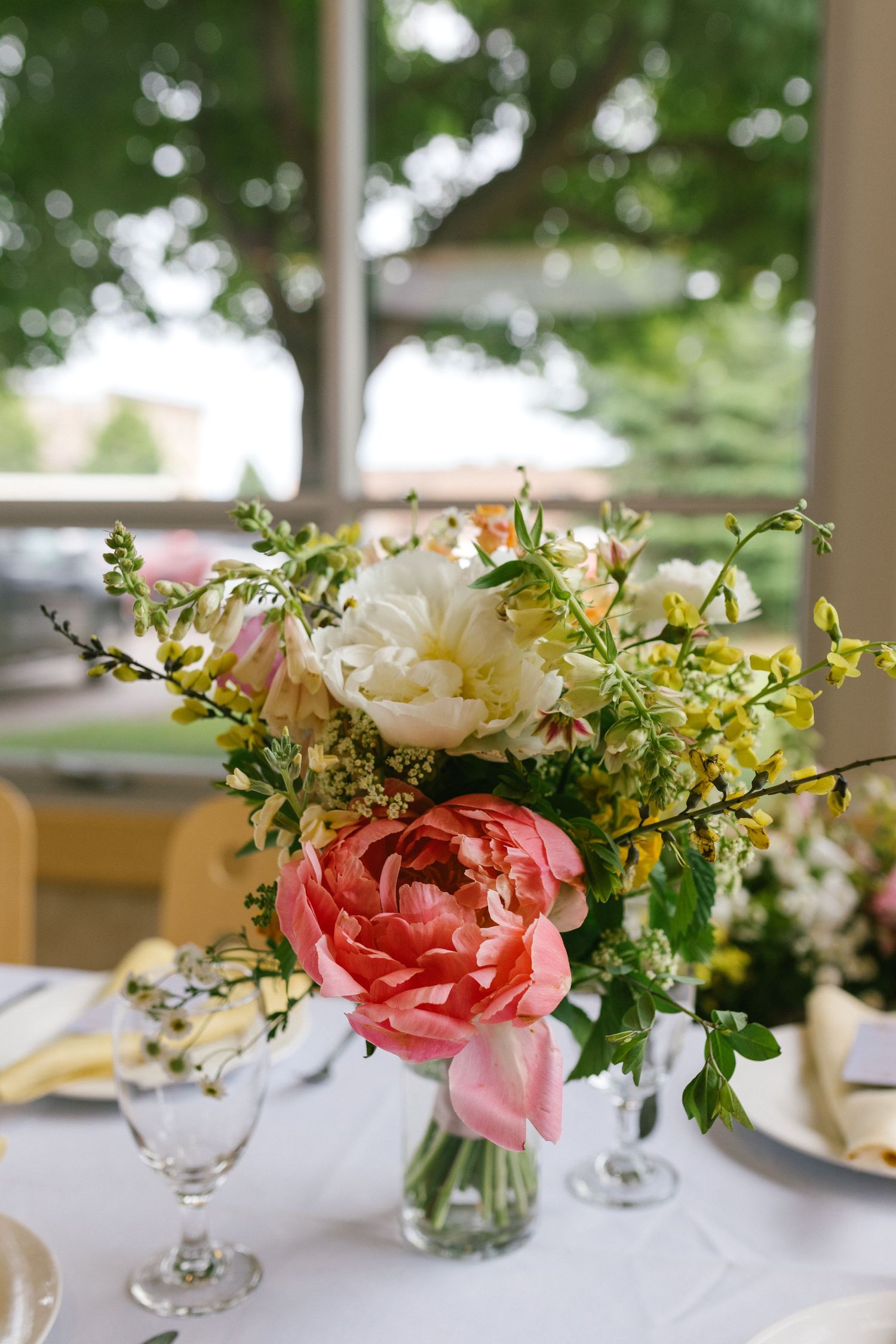 Elegant floral centerpiece with coral and white peonies mixed with yellow wildflowers in a glass vase on white tablecloth.