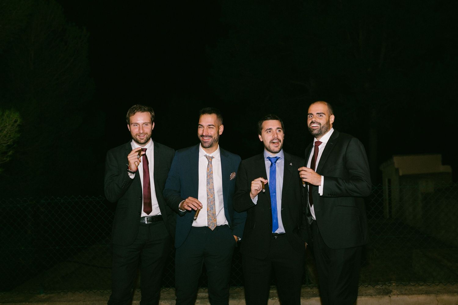 Four men in formal suits and ties stand together smiling at night while having drinks.