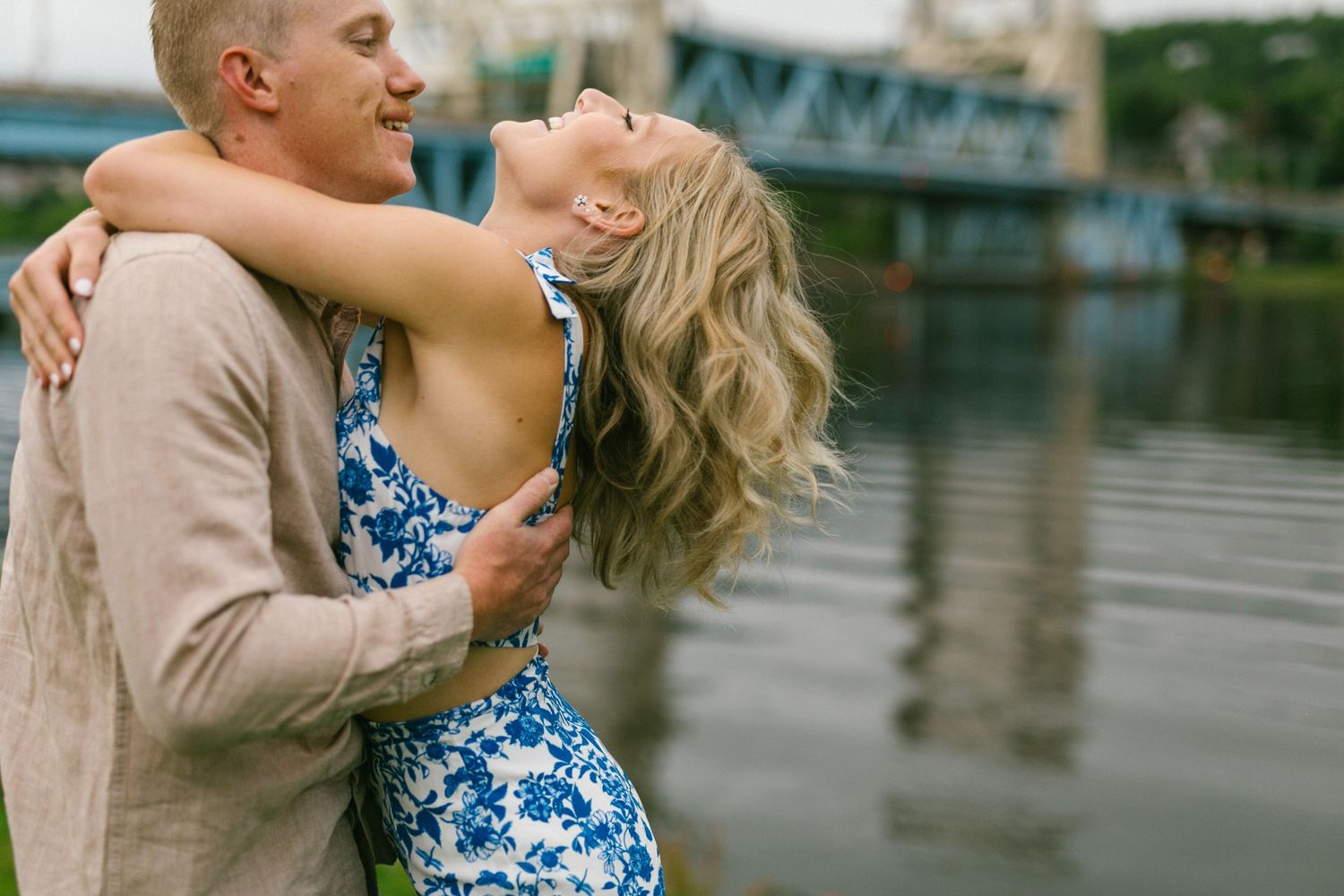 A couple embraces romantically by a waterfront, with the woman wearing a blue and white patterned dress.