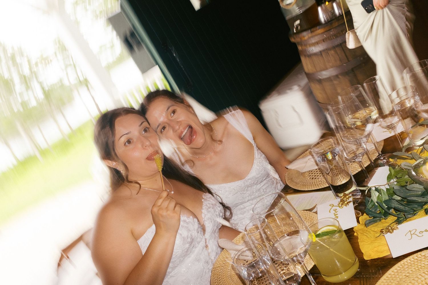 Two women in white dresses smile at a decorated wedding reception table with glassware and greenery.