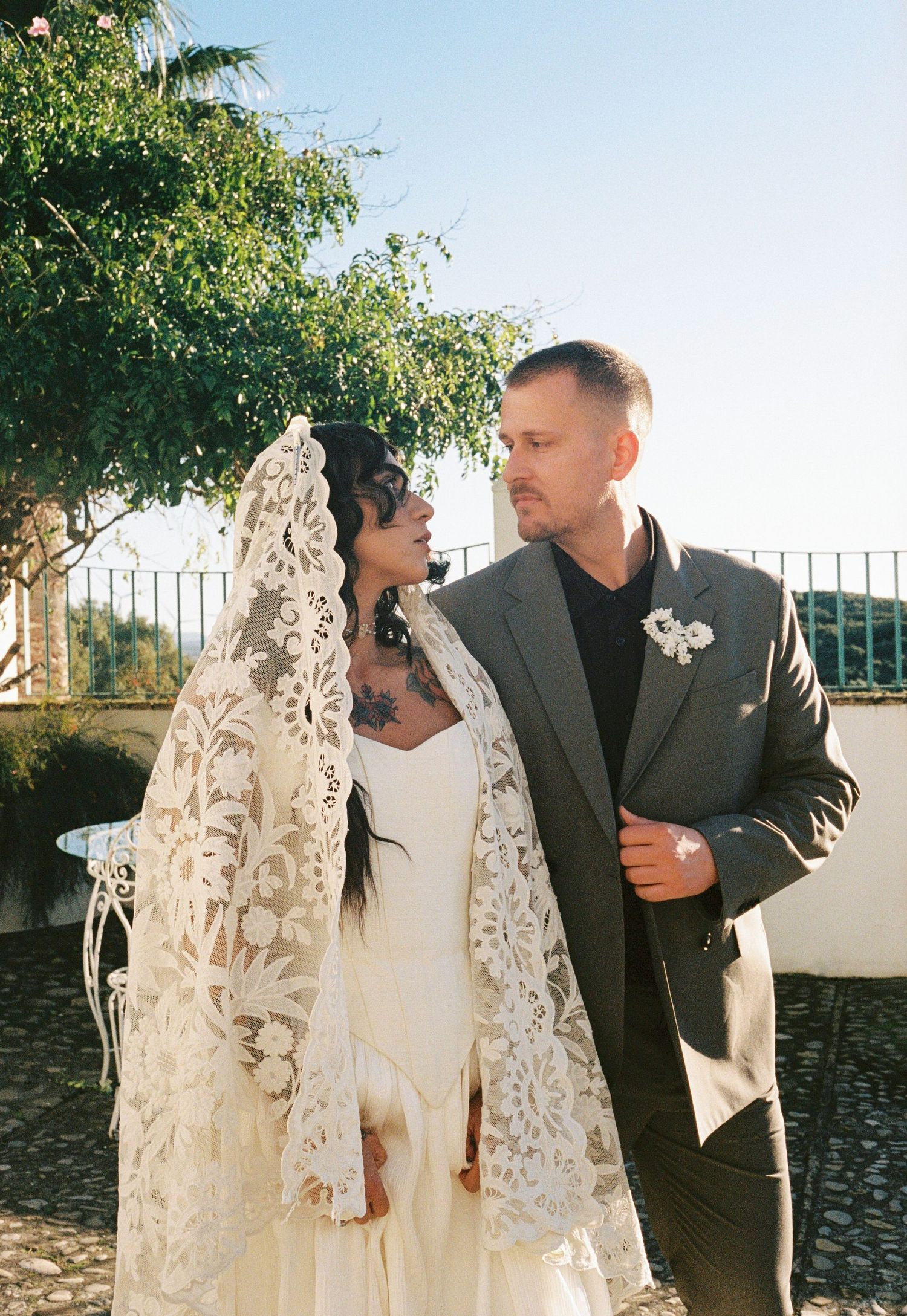Bride in white dress with ornate lace veil gazes at groom in gray suit outdoors.