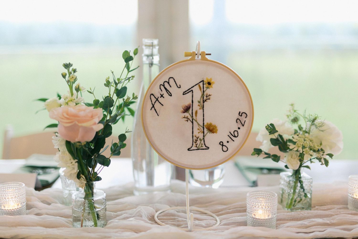 Elegant wedding table settings with embroidery hoops, candles, and delicate floral arrangements in soft natural lighting.
