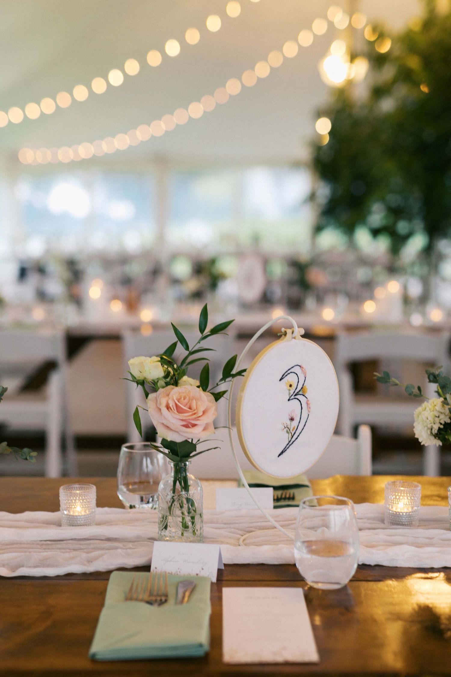 Wedding reception table settings with string lights overhead and delicate floral centerpieces in small vases.