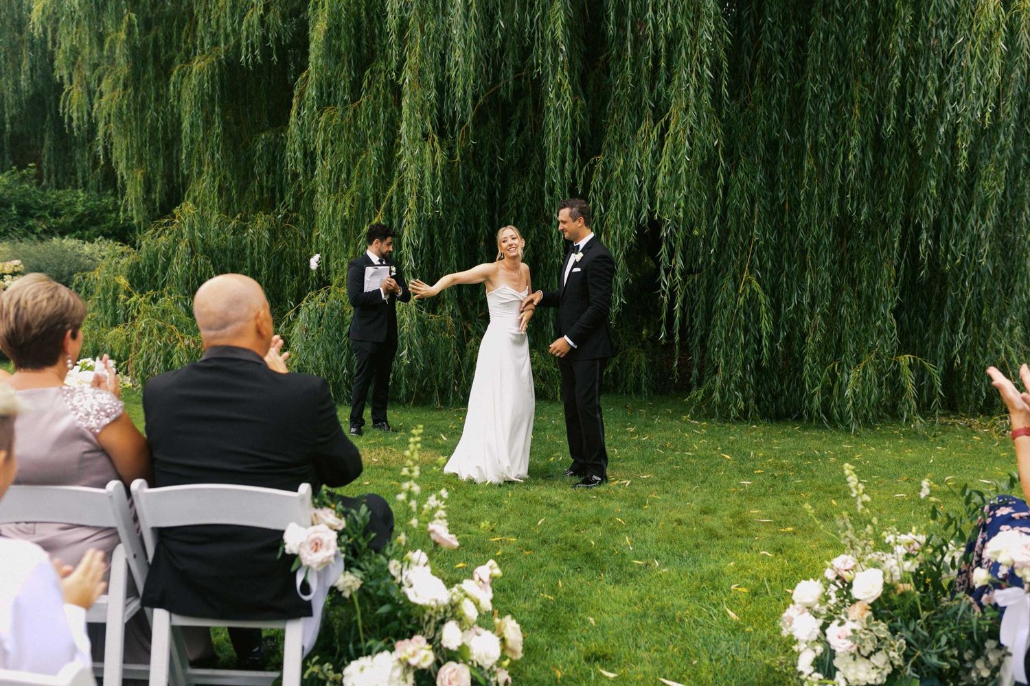 An outdoor wedding ceremony takes place under a majestic weeping willow tree with white floral arrangements and chairs.