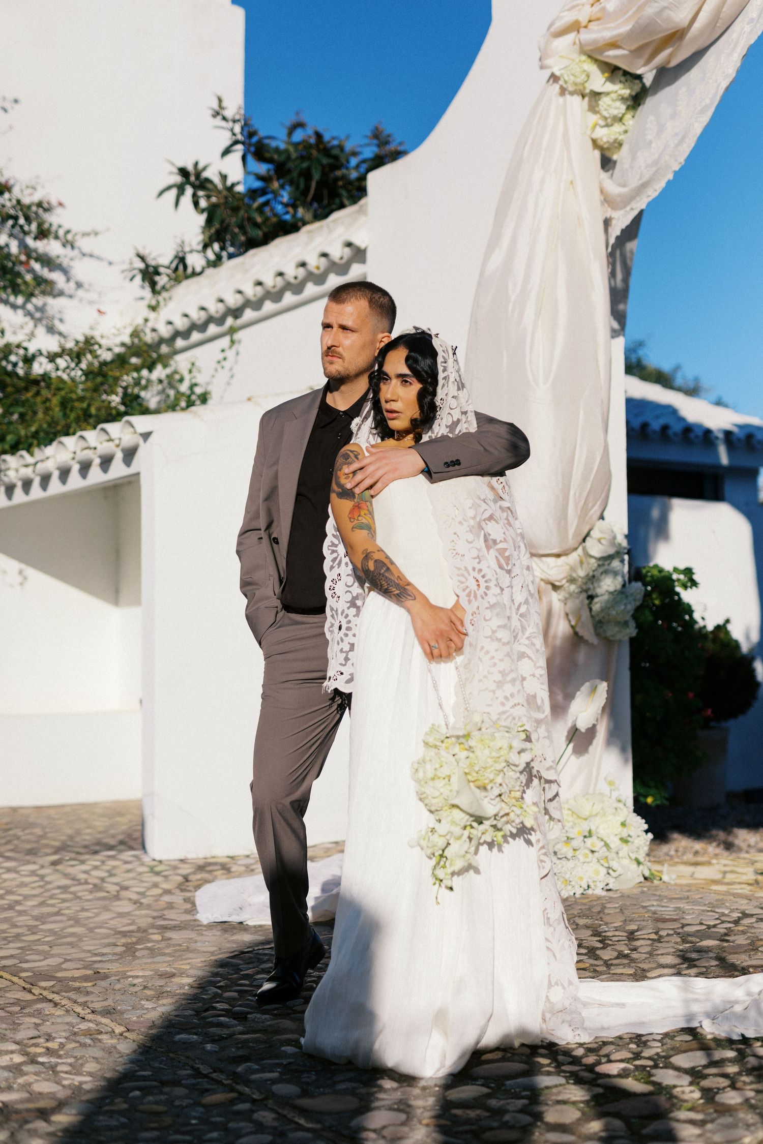 Bride and groom embrace near white arch decorated with flowing fabric and flowers at outdoor wedding venue.