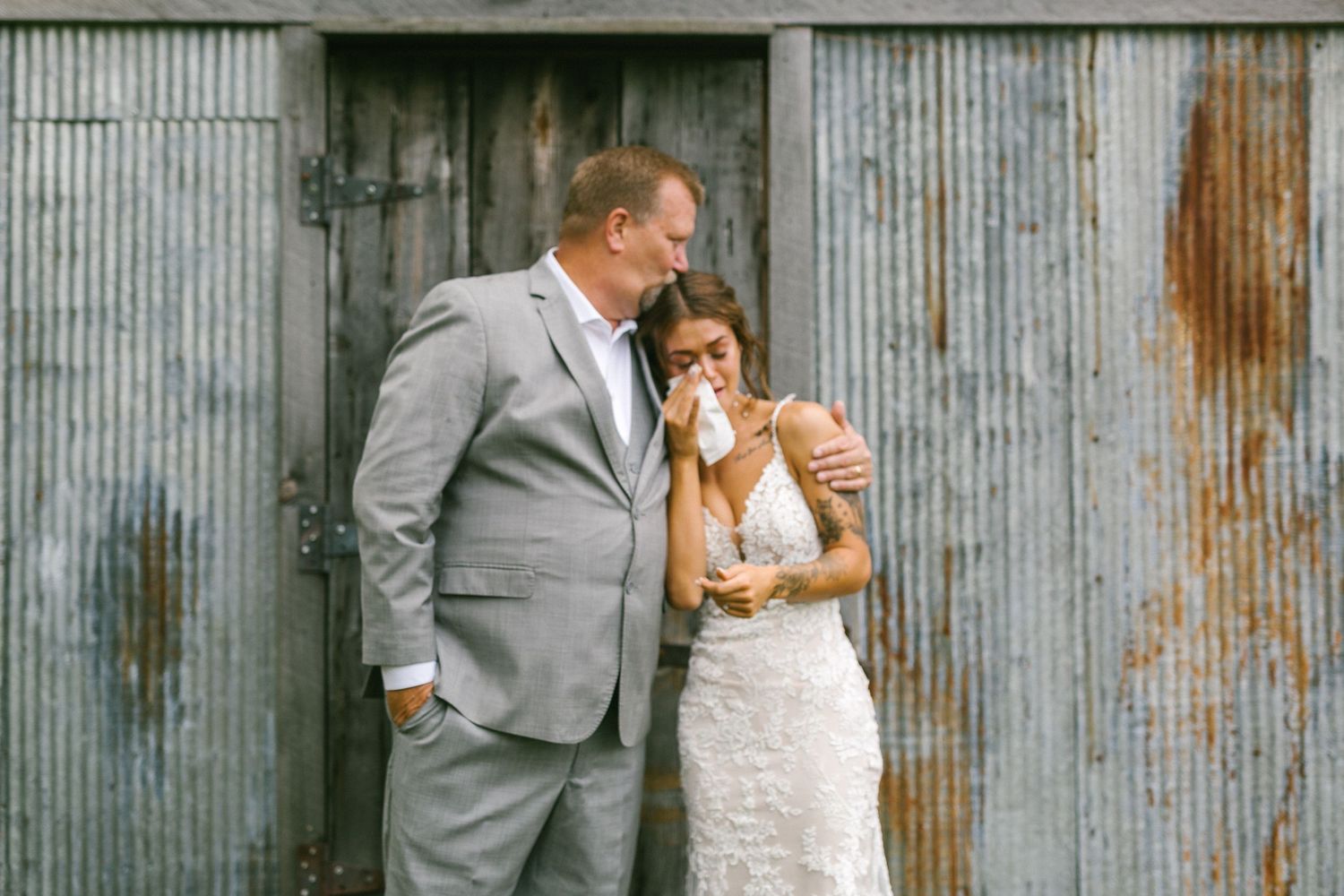 A father and daughter bride in wedding attire share an intimate moment against a rustic corrugated metal wall.