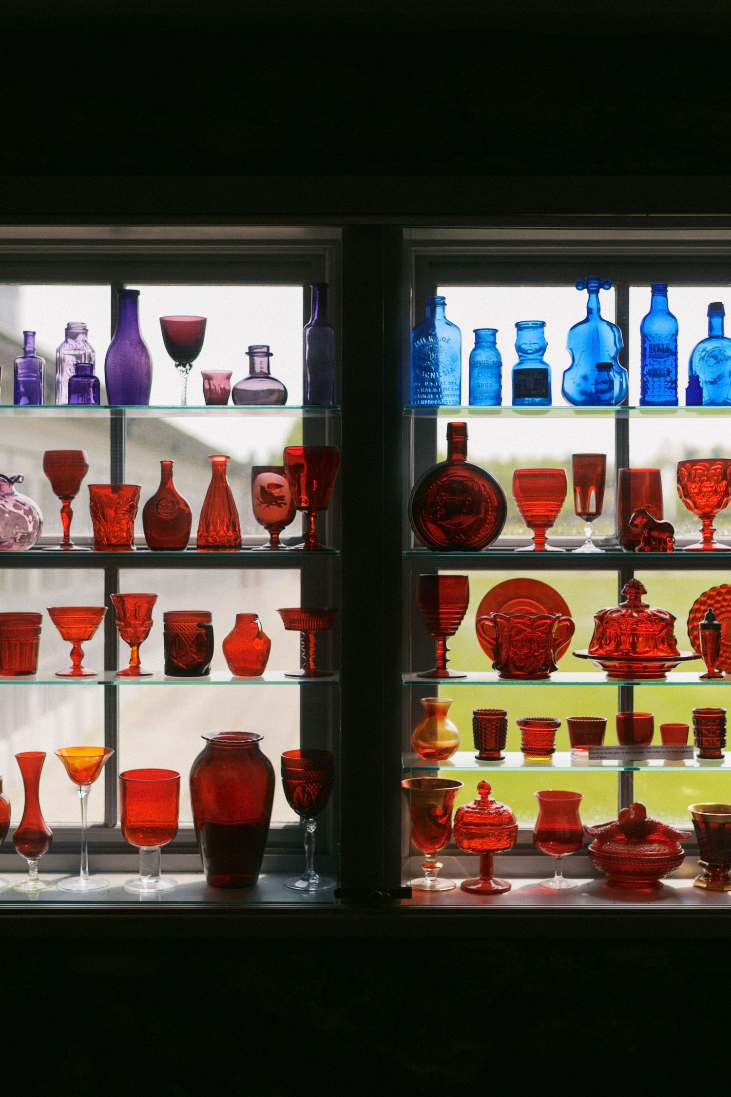 Colorful glass display featuring red and blue vintage bottles and vases illuminated on backlit shelves.